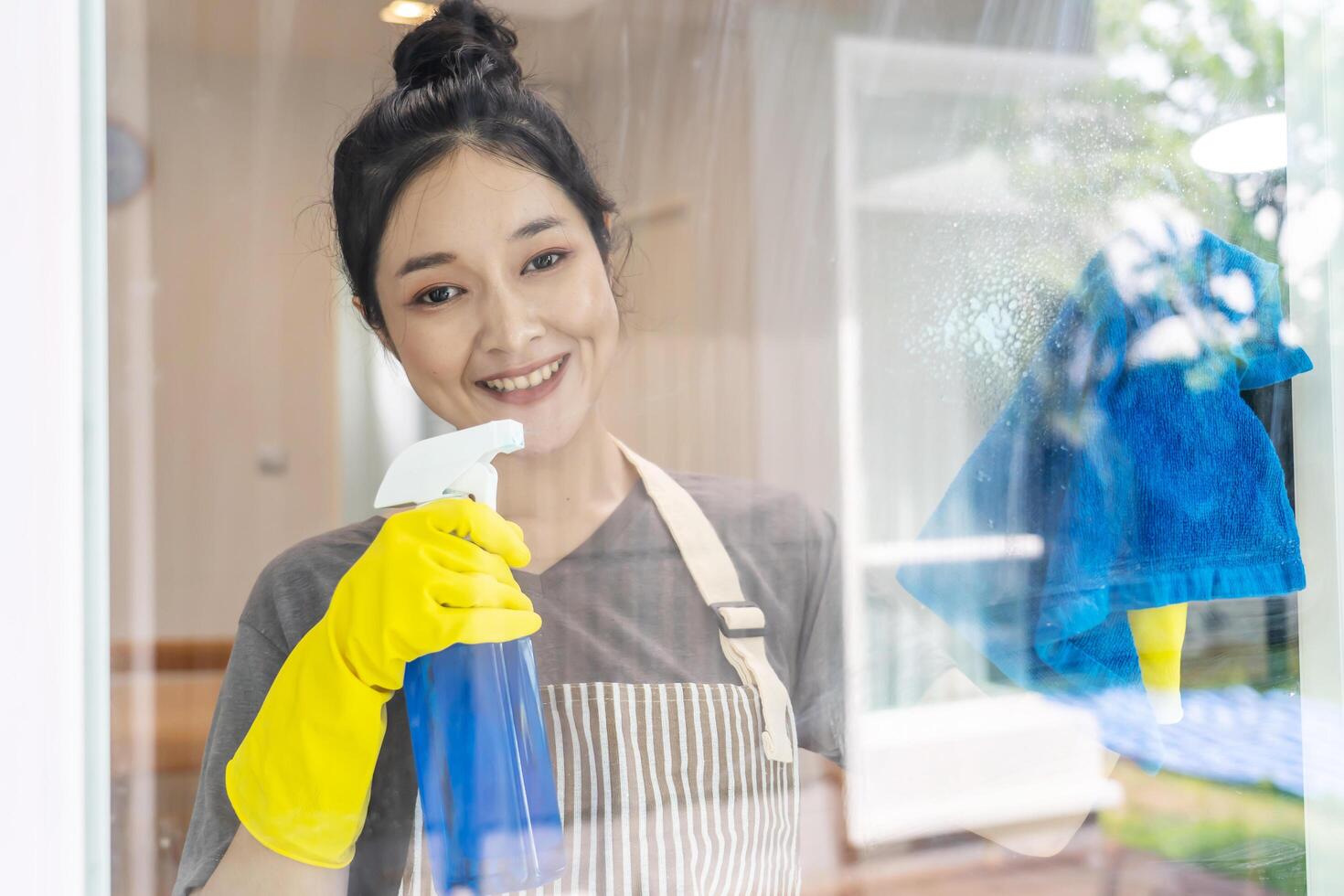 Portrait of cheerful positive young woman asian in yellow protective rubber gloves cleaning window with rag and cleanser spray at home, enjoying housework, happily performing domestic chores photo