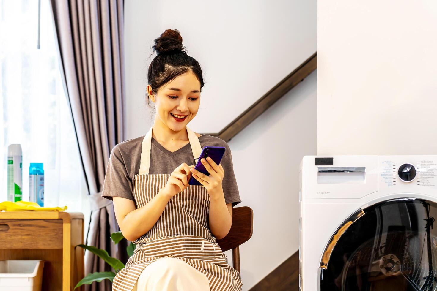 Young asian woman are playing with mobile phone in home sitting near the washing machine, while waiting for the washing machine to finish working photo