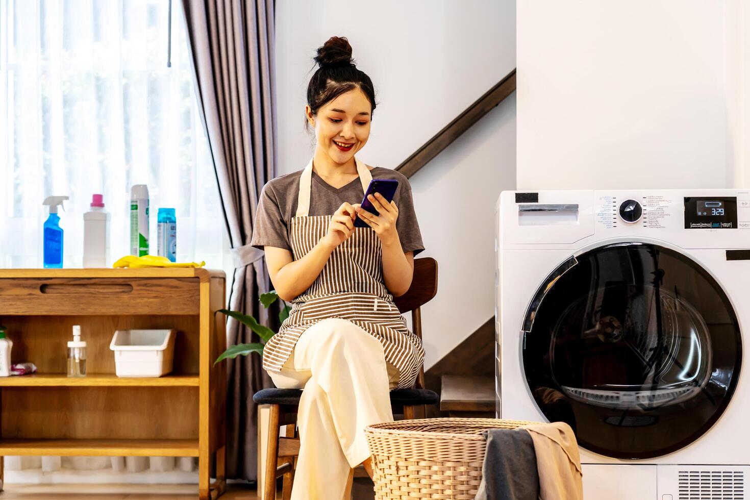 Young asian woman are playing with mobile phone in home sitting near the washing machine, while waiting for the washing machine to finish working photo