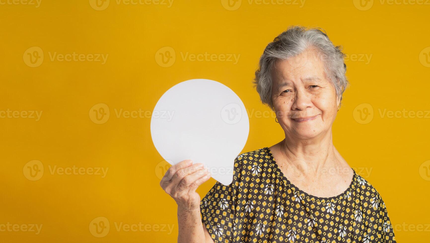 An elderly Asian woman with short gray hair holding a blank speech bubble and looking at the camera with a smile while standing on a yellow background in the studio photo