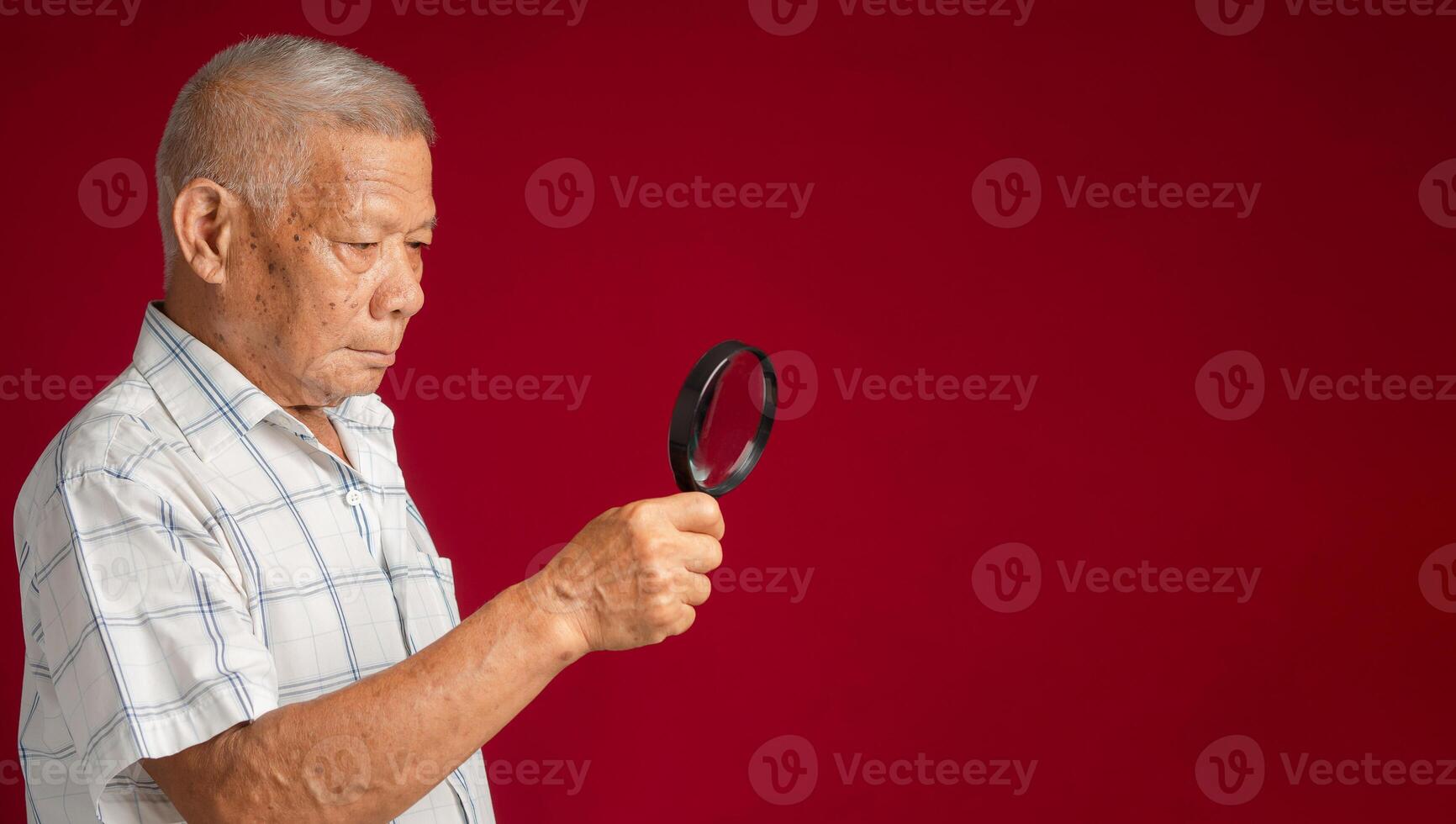 Senior man in a white shirt holding while standing a magnifying glass over a red background in the studio. Aged people and search concept photo