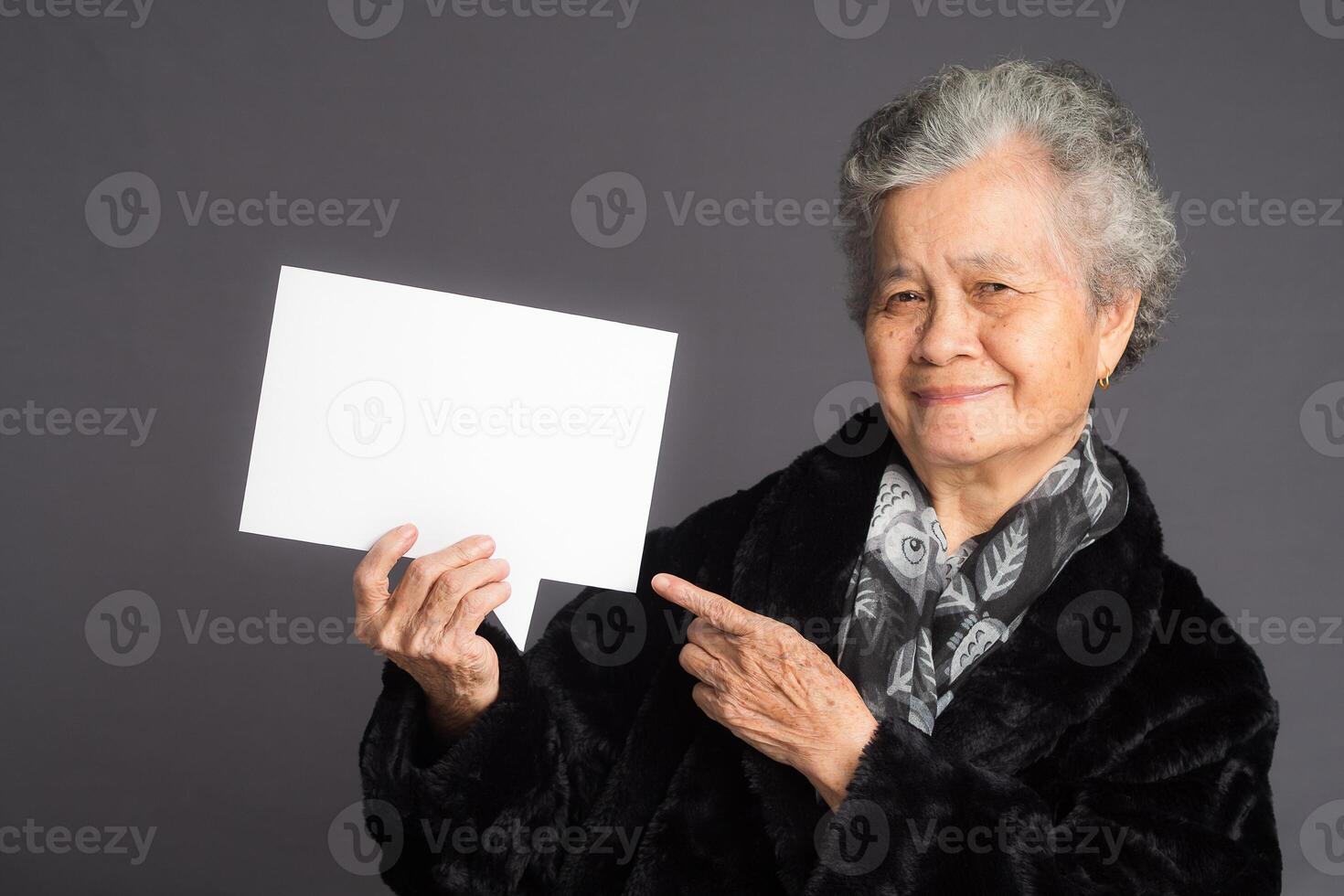 An elderly Asian woman in a sweater with short gray hair holding a blank speech bubble and looking at the camera with a smile while standing on a gray background photo