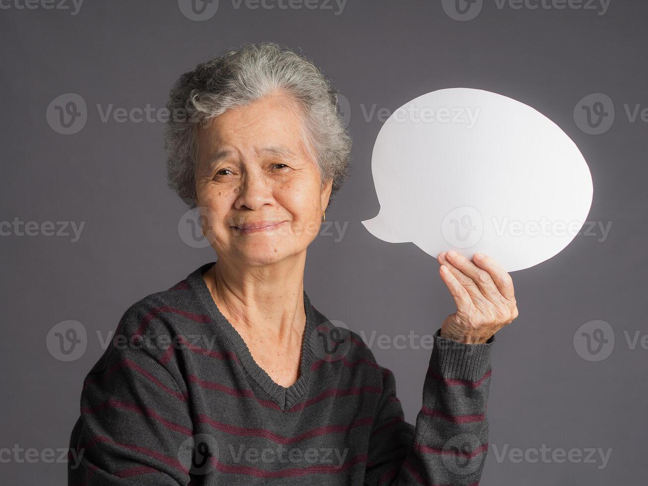 An elderly Asian woman in a sweater with short gray hair holding a blank speech bubble and looking at the camera with a smile while standing on a gray background photo