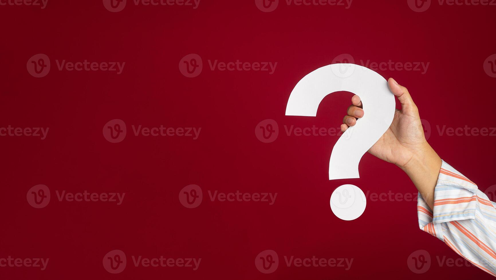 Hand of a young man in a shirt holding a white question mark paper while standing with a red background in the studio photo