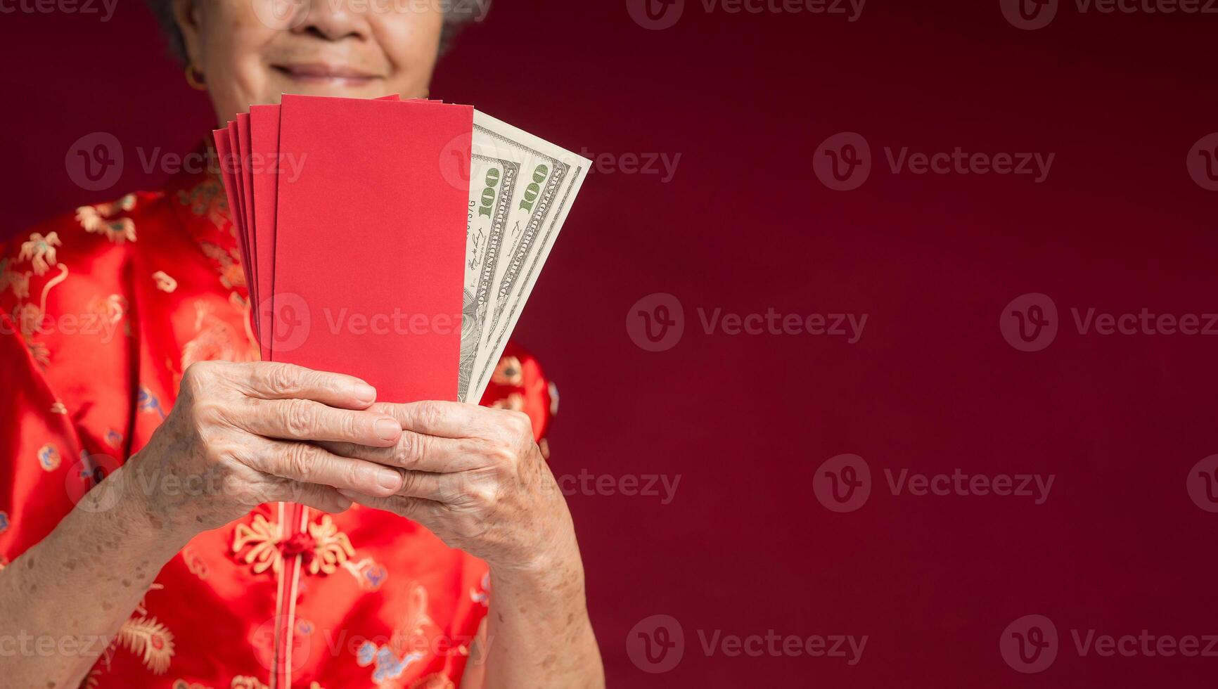 An elderly Asian woman wearing a traditional cheongsam qipao dress holding Angpao while standing over a red background photo