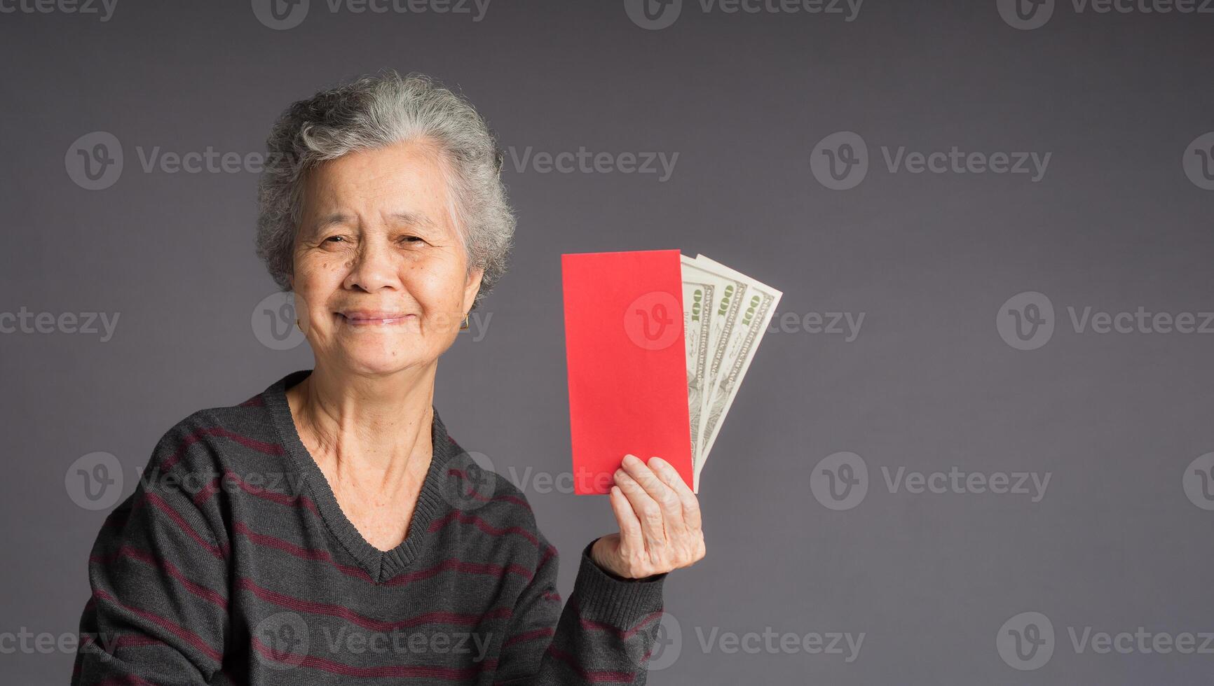 An elderly Asian woman in a sweater holding Angpao with US dollar banknote and looking at the camera with a smile while standing on gray background photo