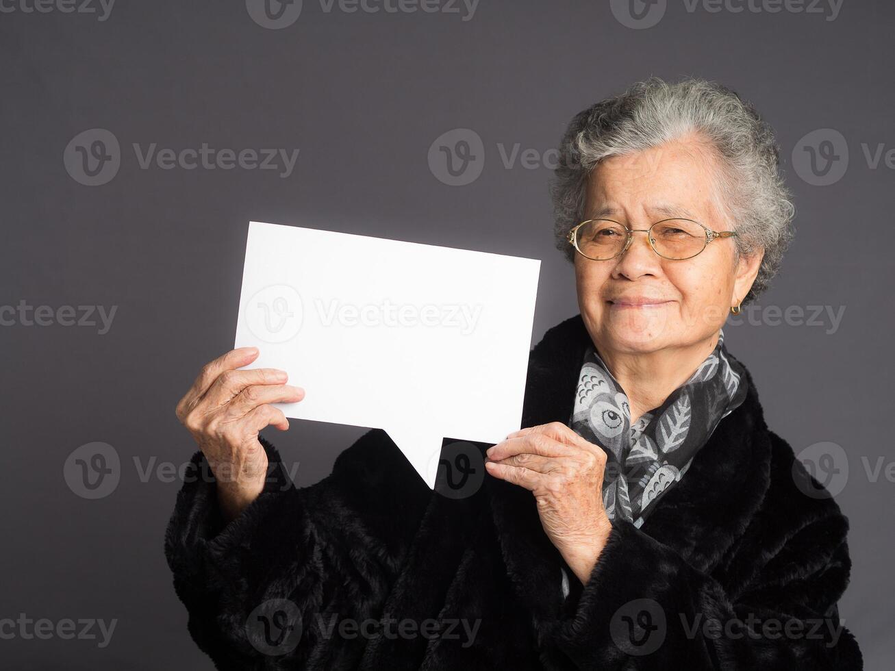 An elderly Asian woman in a sweater with short gray hair holding a blank speech bubble and looking at the camera with a smile while standing on a gray background photo