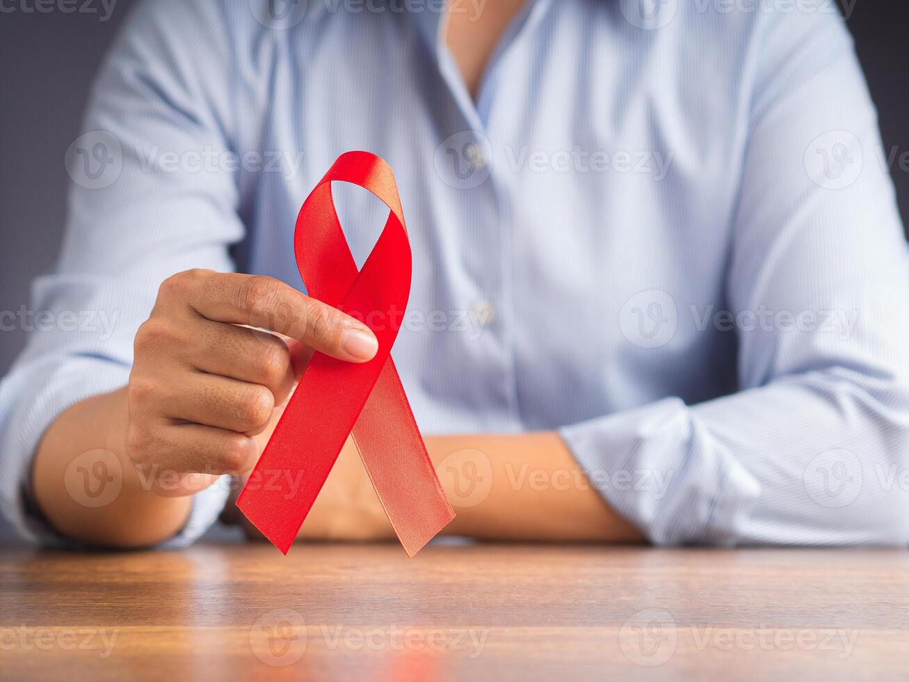 Red ribbon awareness concept. Woman in a blue shirt with hand holding a red ribbon while sitting in the office. World AIDS day, World cancer day, World hypertension day. Space for text photo