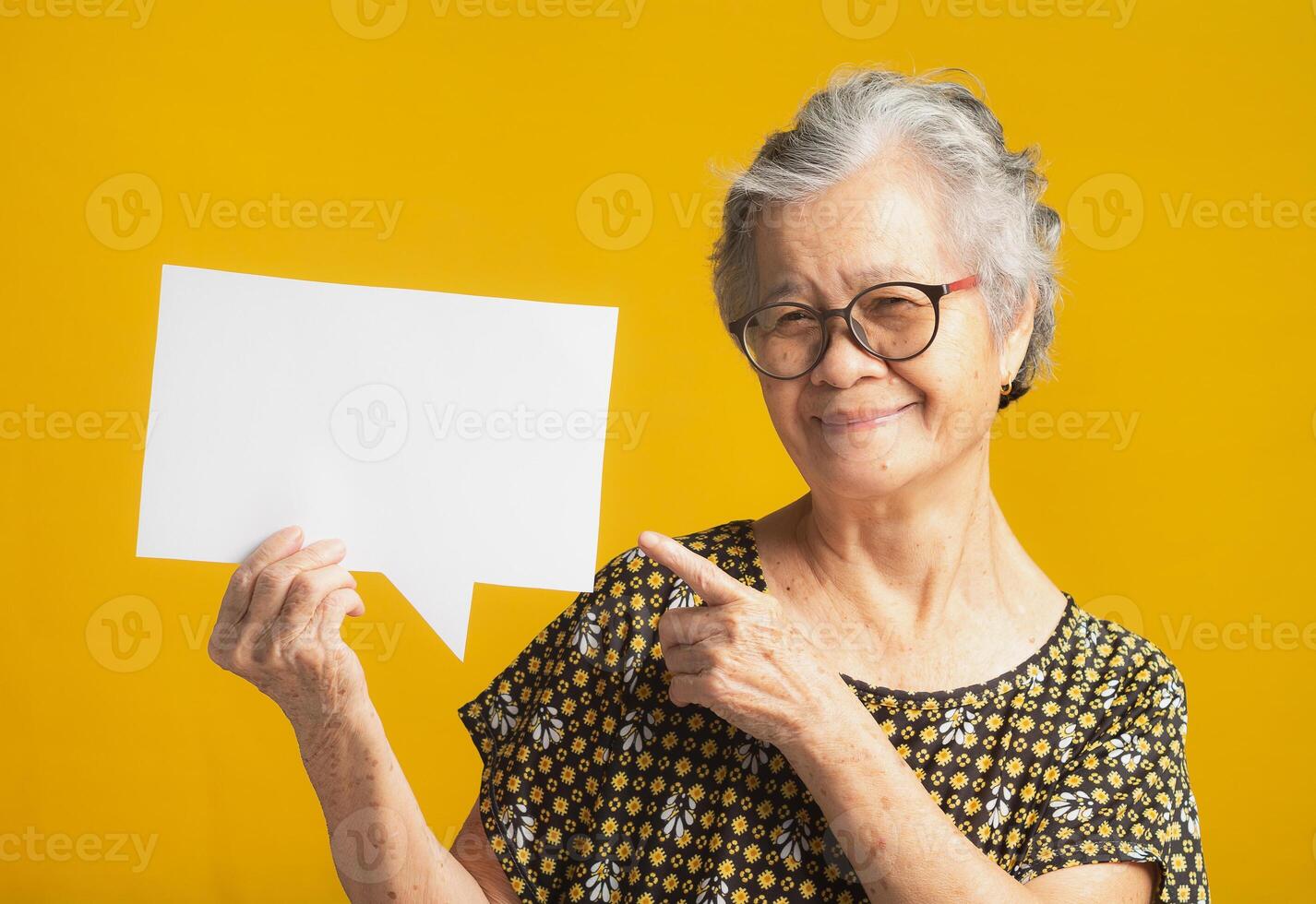 Speech bubble concept. An elderly Asian woman with short gray hair wearing glasses, holding and pointing at a blank paper bubble balloon with a smile while standing on a yellow background photo