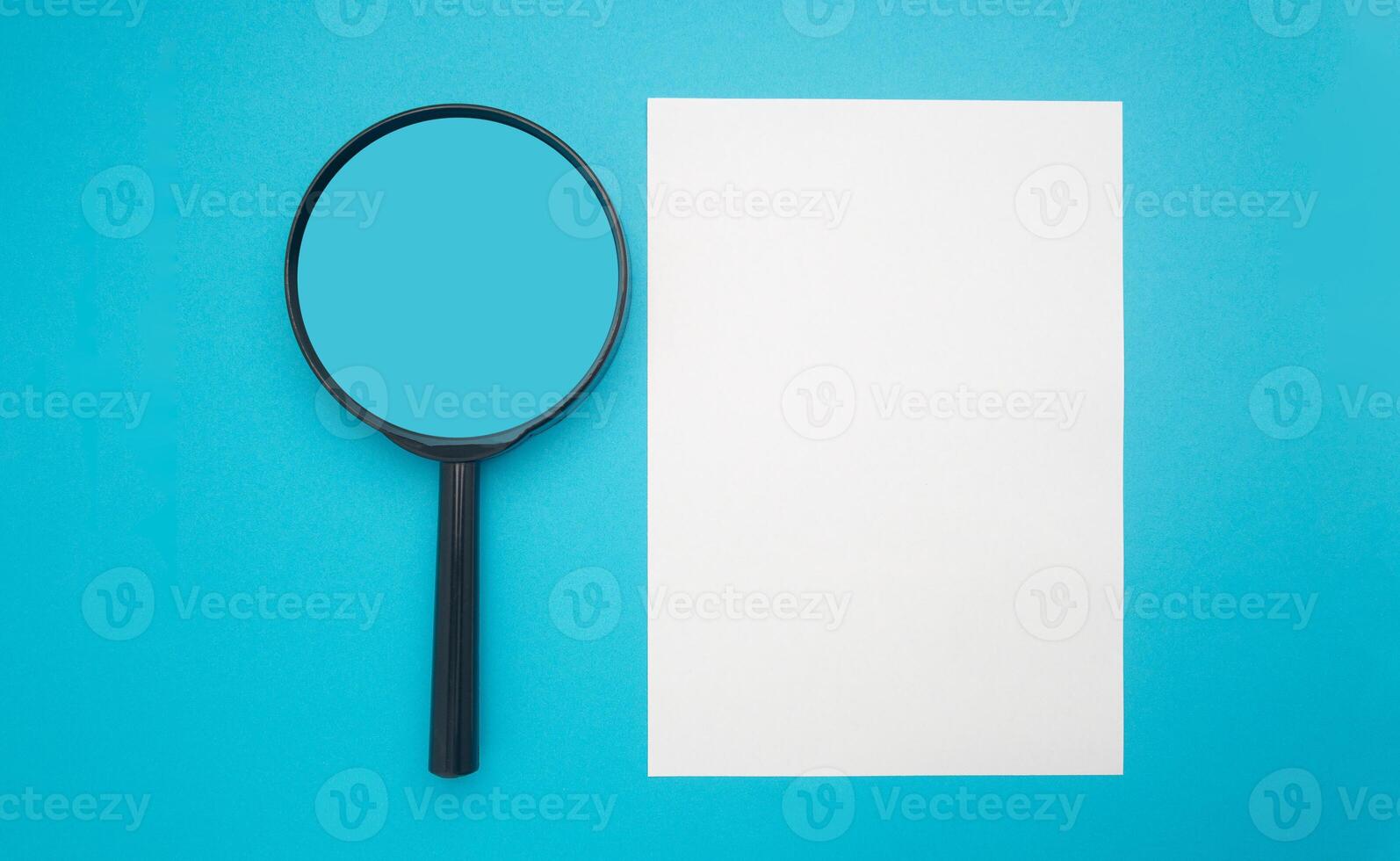 Top view of a magnifying glass and a blank white paper isolated on blue background. Search and document concept photo