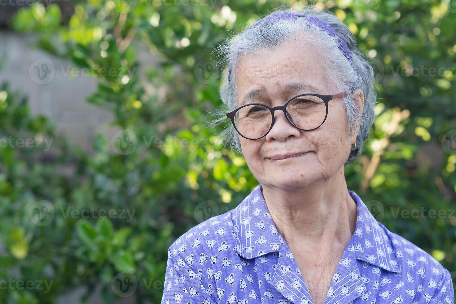 Portrait of an elderly Asian woman with short gray hair, wearing glasses, and looking away with a smile while standing outdoor. Aged people and relaxation concept photo