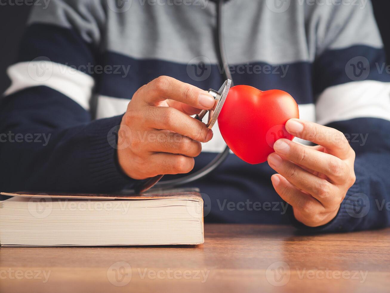 Midsection of a doctor in a sweater holding a stethoscope checking a red heart shape while sitting in the hospital or clinic. Close-up photo. Space for text. Medical, treatment, and healthcare concept photo