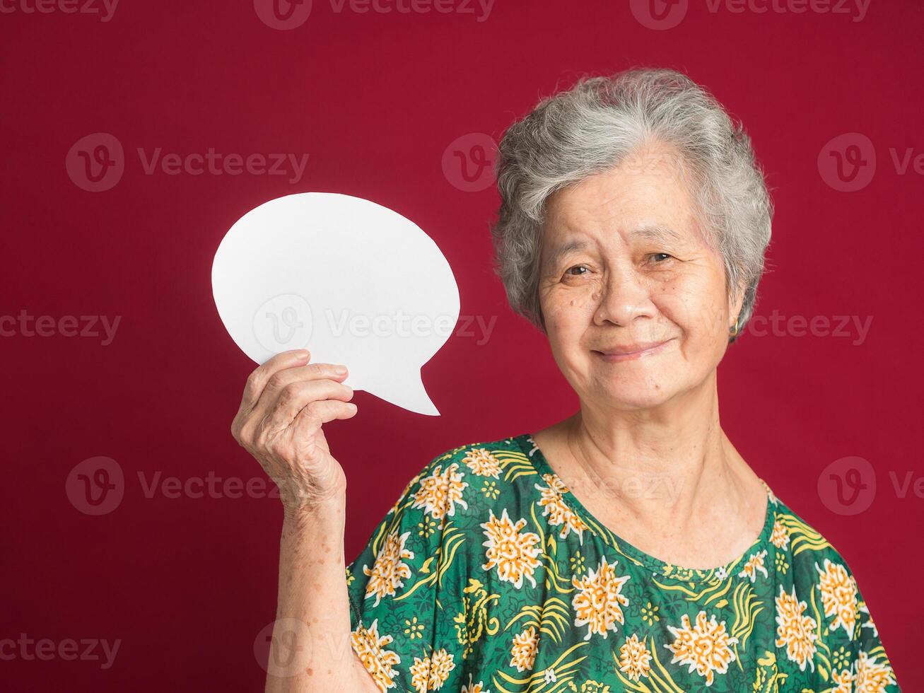 Concept of a speech bubble. Happy senior woman with short gray hair holding a blank paper and looking at the camera with a smile while standing on a red background in the studio photo