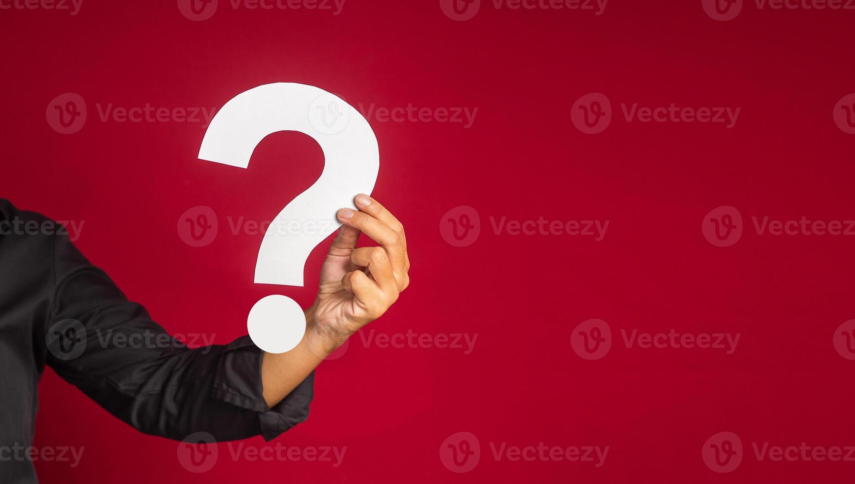 Question mark concept. Hand of a businessman in a black shirt holding a white question mark while standing with a red background in the studio. Close-up photo. Space for text photo