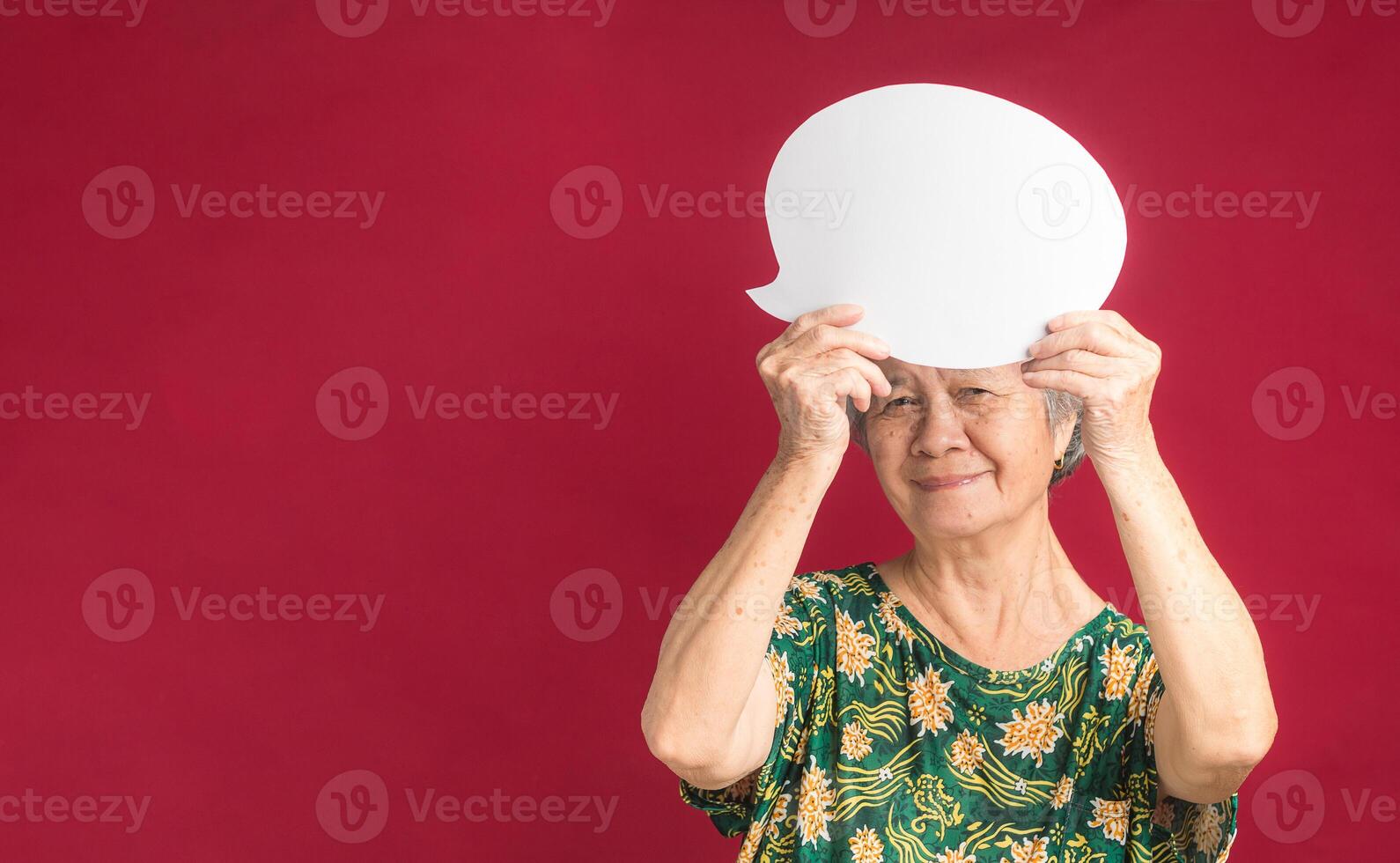 A speech bubble concept. Happy senior woman with short gray hair holding a blank paper bubble balloon and looking at the camera with a smile while standing on a red background in the studio photo