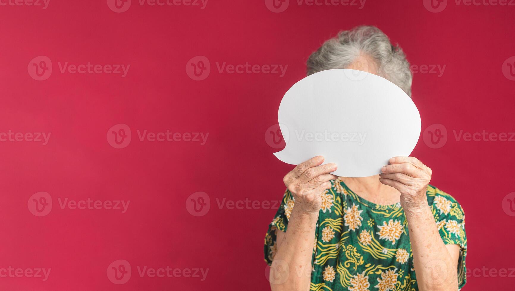 Concept of a speech bubble. A senior woman with short gray hair holding a blank white paper speech bubble closed her face while standing on a red background in the studio. Space for text photo