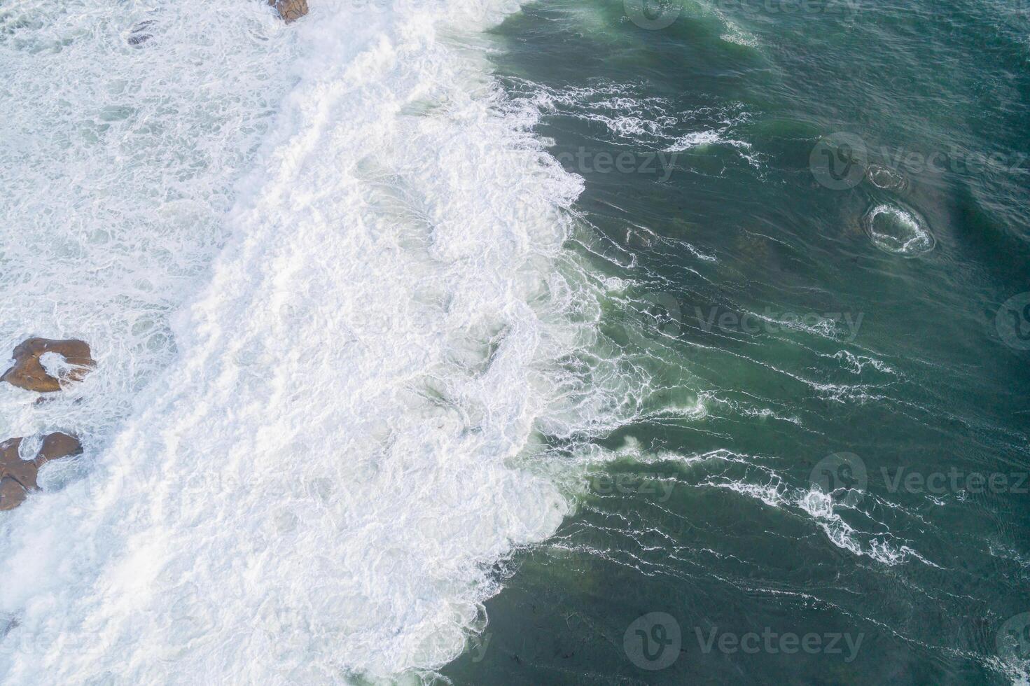 Aerial photo of a rocky shore with crashing waves creating foam.