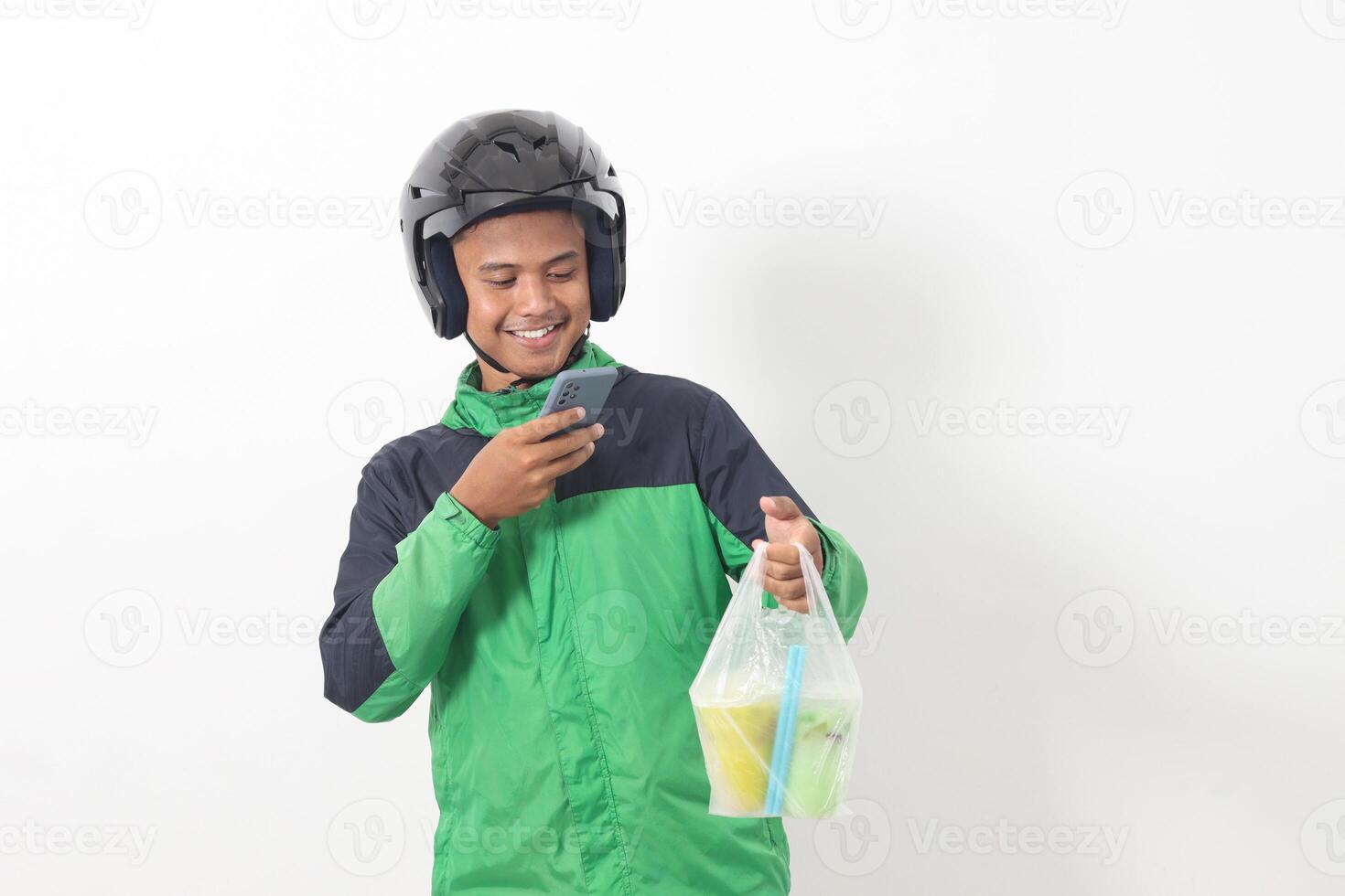 Portrait of Asian online taxi driver wearing green jacket and helmet delivering the beverages in plastic cup to customer, while using mobile phone. Isolated image on white background photo