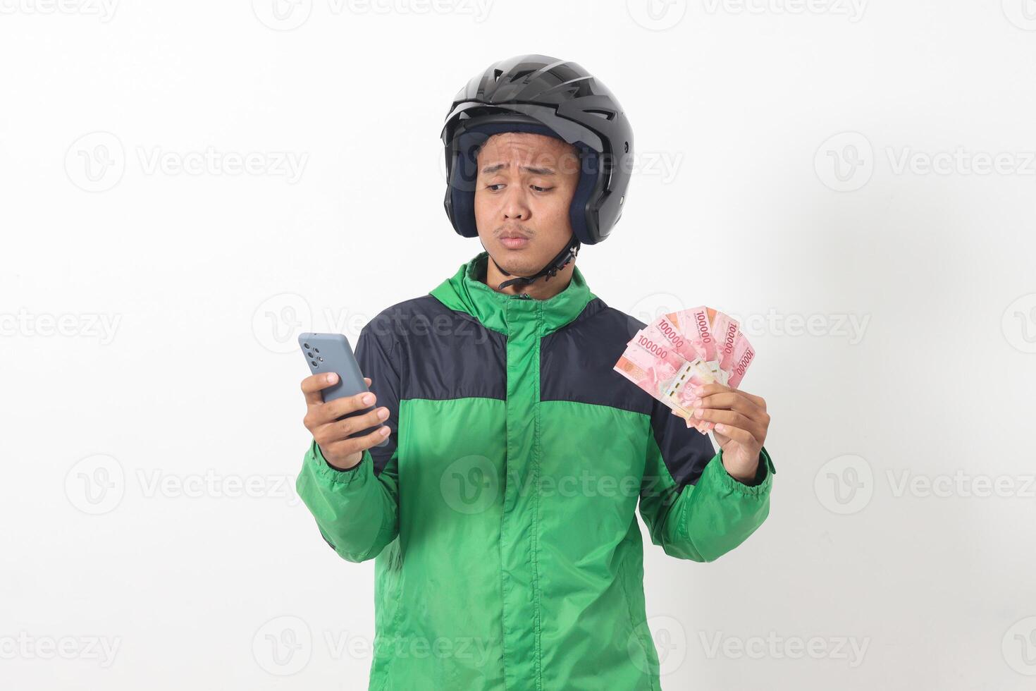 Portrait of Asian online taxi driver wearing green standing against white background, smiling and looking at camera, holding a bunch of rupiah money photo