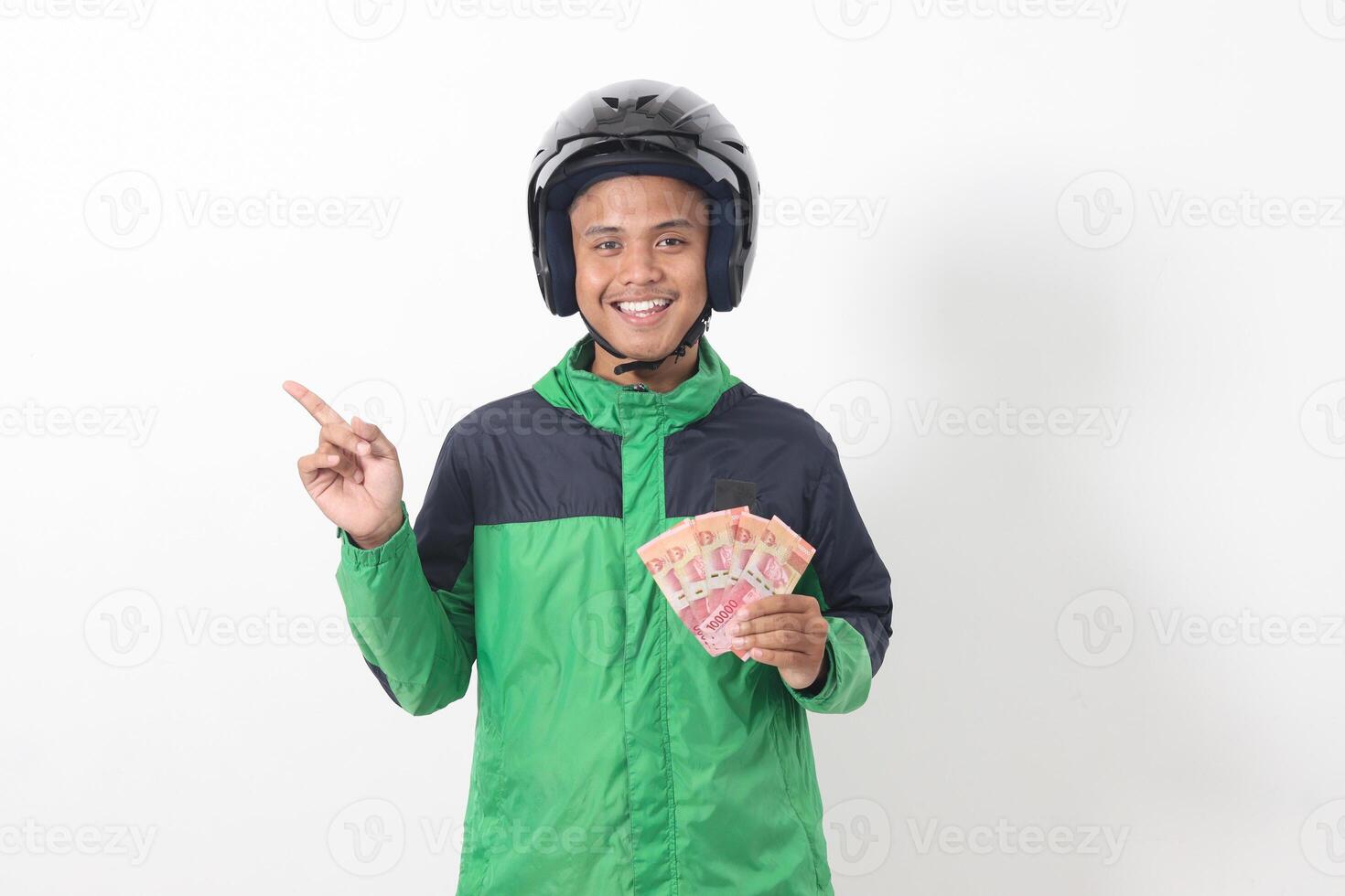 Portrait of Asian online taxi driver wearing green standing against white background, holding a bunch of rupiah money while pointing to the side photo