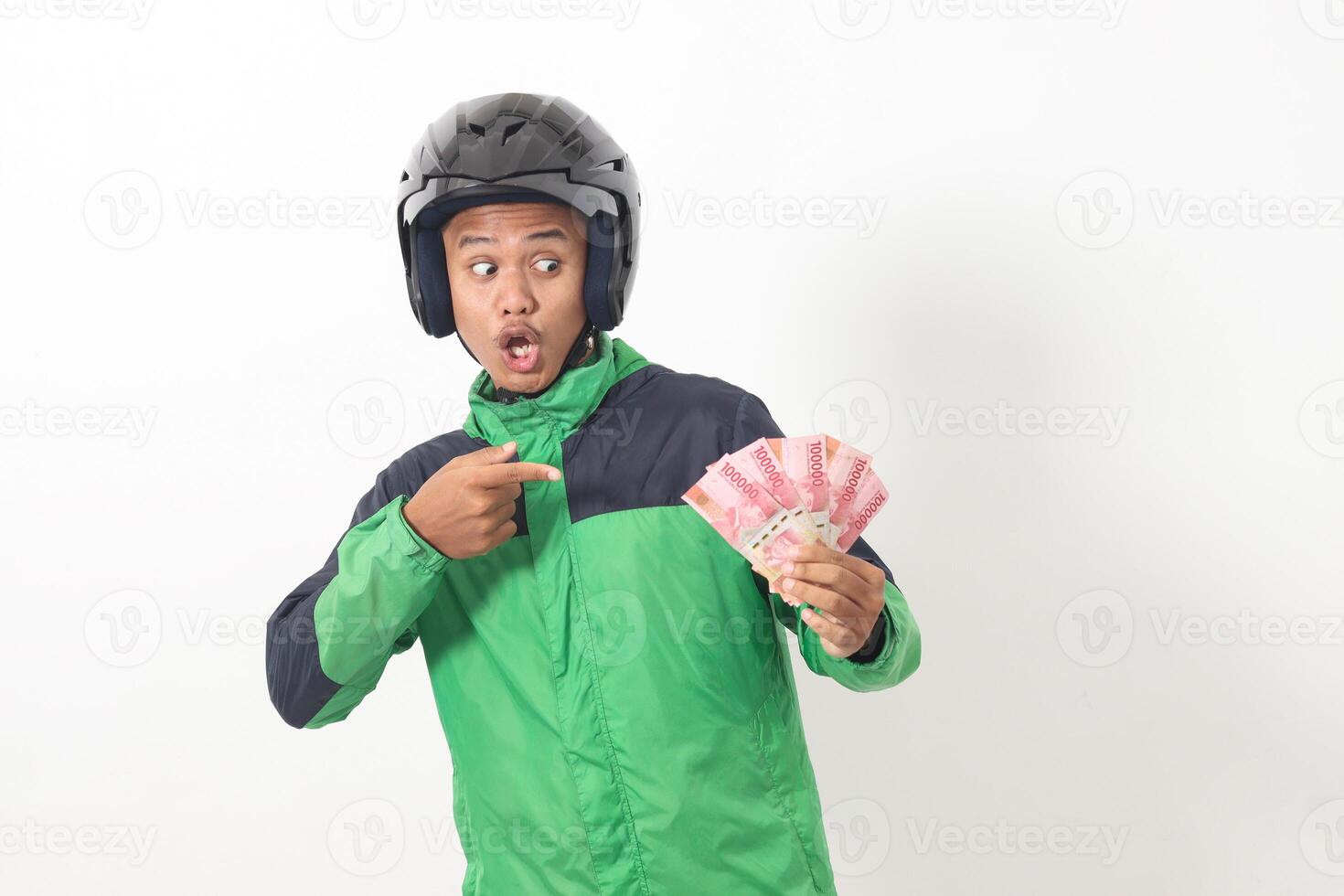 Portrait of Asian online taxi driver wearing green standing against white background, holding a bunch of rupiah money while pointing to the side photo