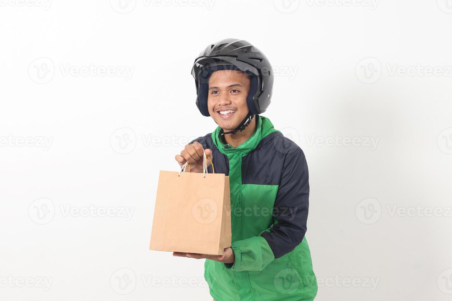 Portrait of Asian online courier driver wearing green jacket and helmet giving craft paper shopping bag with gift inside. Isolated image on gray background photo