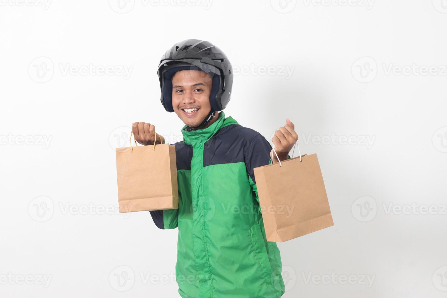 Portrait of Asian online courier driver wearing green jacket and helmet giving craft paper shopping bag with gift inside. Isolated image on gray background photo