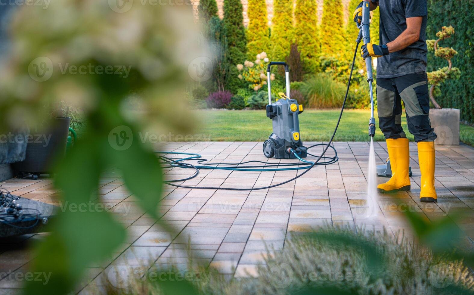 Man Cleaning Patio Tiles With a Pressure Washer in a Well-Maintained Garden on a Sunny Day photo