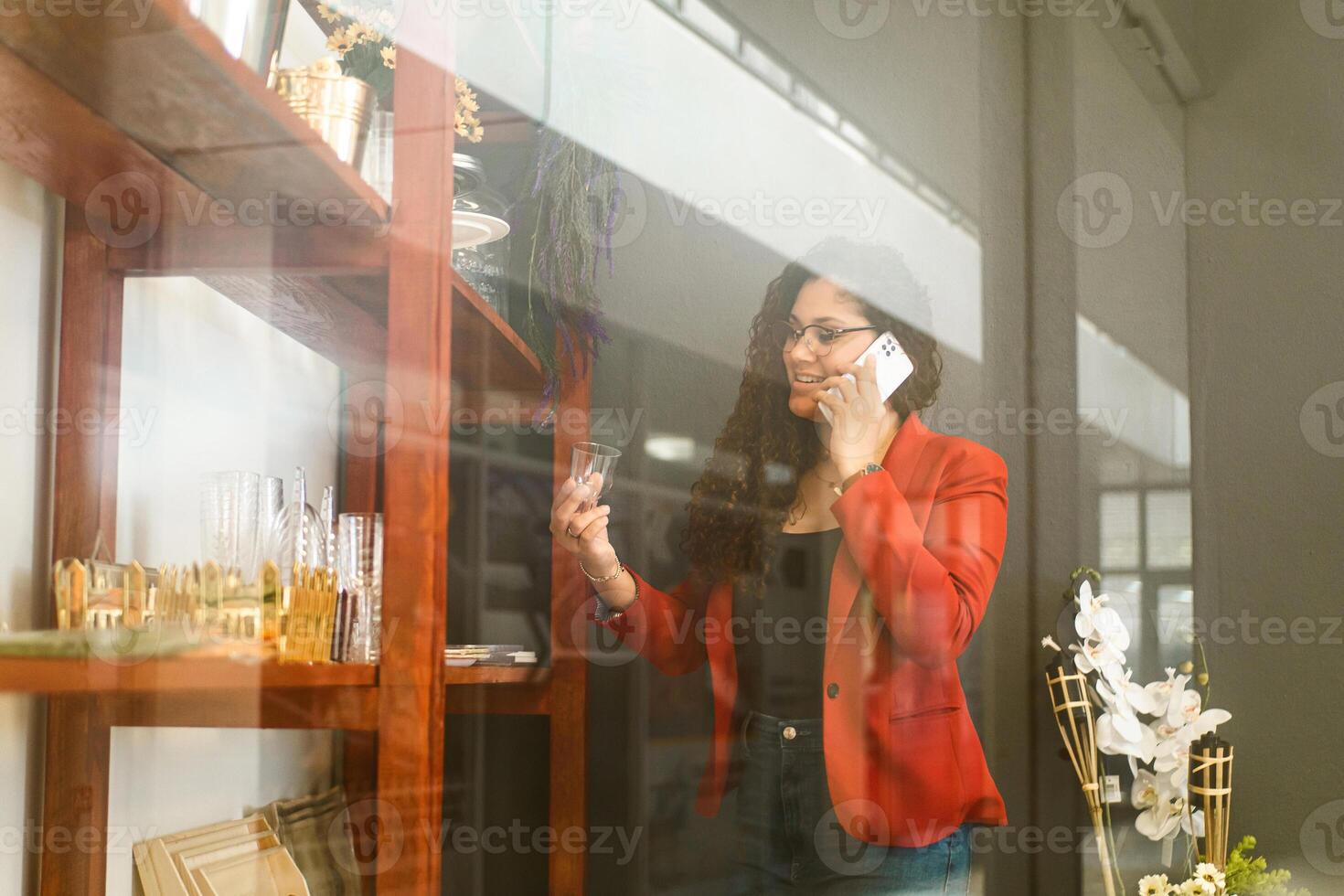 Latin American businesswoman holding a glass from a shelf of items and making a phone call with a white mobile. photo