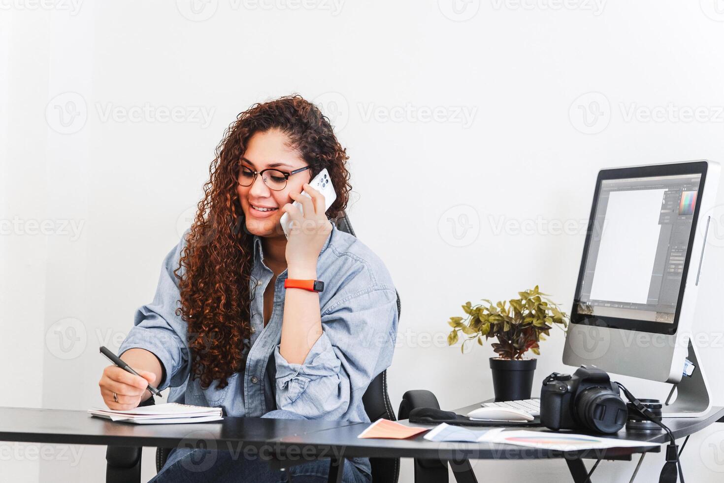 Young Latin American woman photographer and entrepreneur making notes in an agenda while making a call with her mobile phone photo