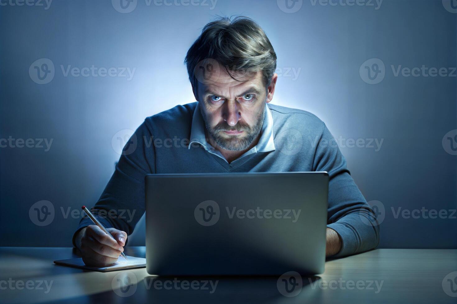 A man with a beard and a white shirt is sitting at a table with a laptop and a cup of coffee. He looks angry and is staring at the laptop screen. photo