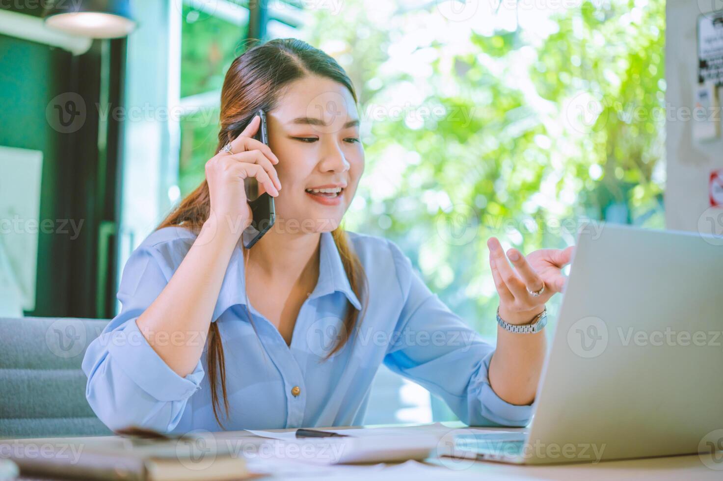 Business asian woman sitting at her desk with laptop using mobile phone chatting with customers or giving financial advice. photo
