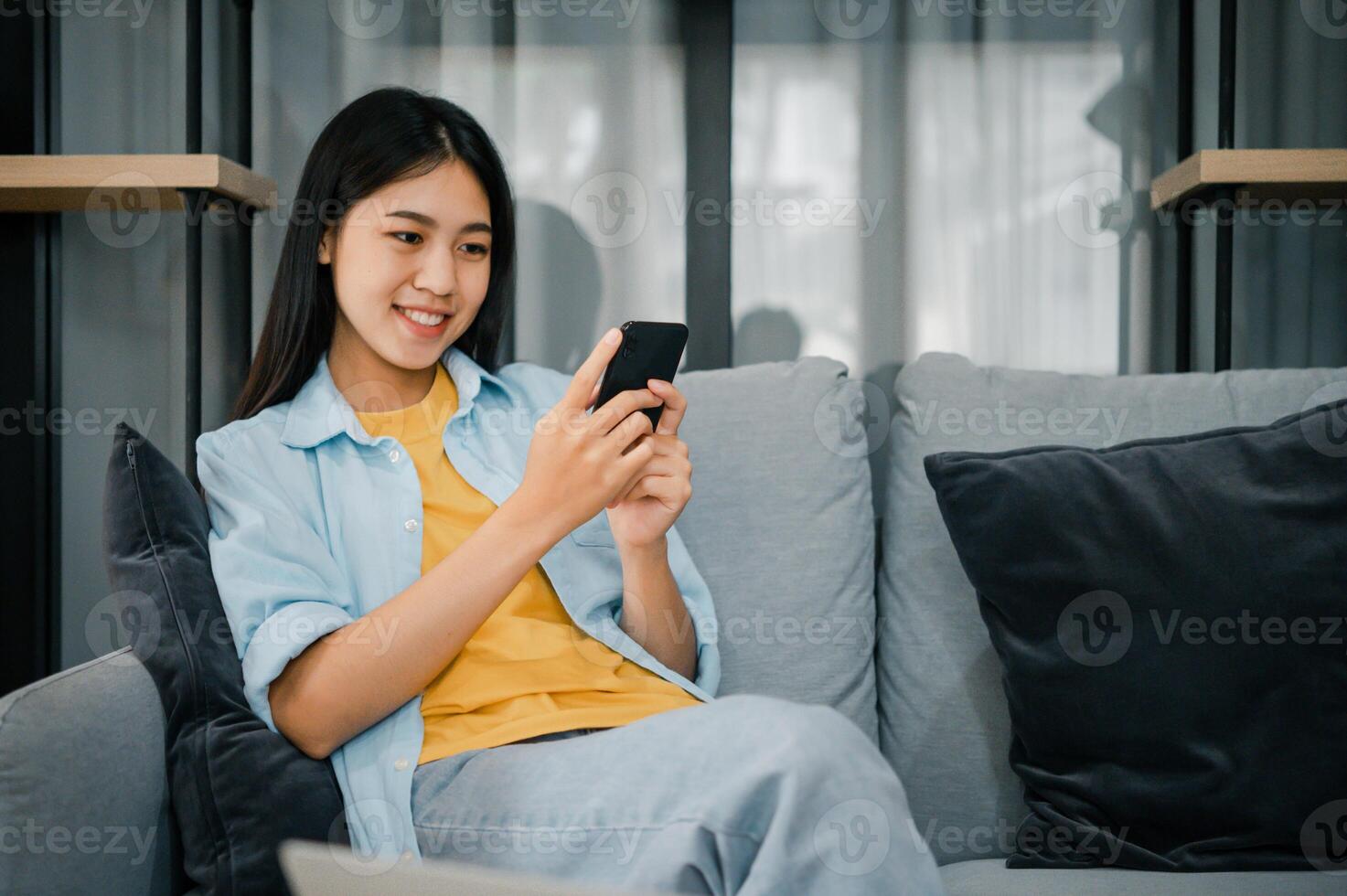 Smiling young asian woman using mobile phone checking social media,app playing game, shopping online, ordering delivery relax while sitting on a couch at home with laptop computer photo