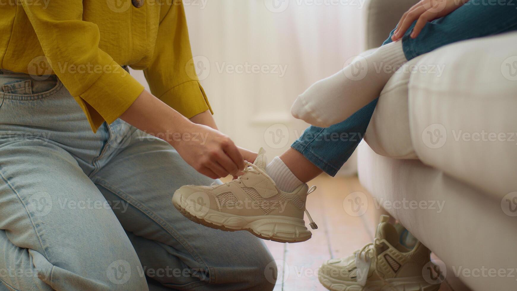 Unrecognizable loving Caucasian woman mother carer female hands lacing ties shoelaces unknown little girl daughter helping tying sneakers child kid in living room at morning. Upbringing family support photo