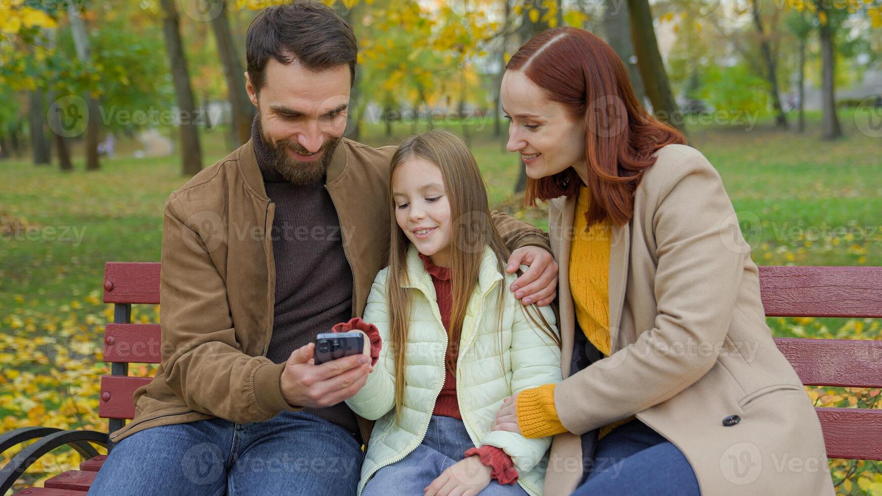 Caucasian happy smiling family relax weekend holiday on city bench autumn park using mobile phone together smile app shopping online smartphone talking little daughter girl child kid mother father photo
