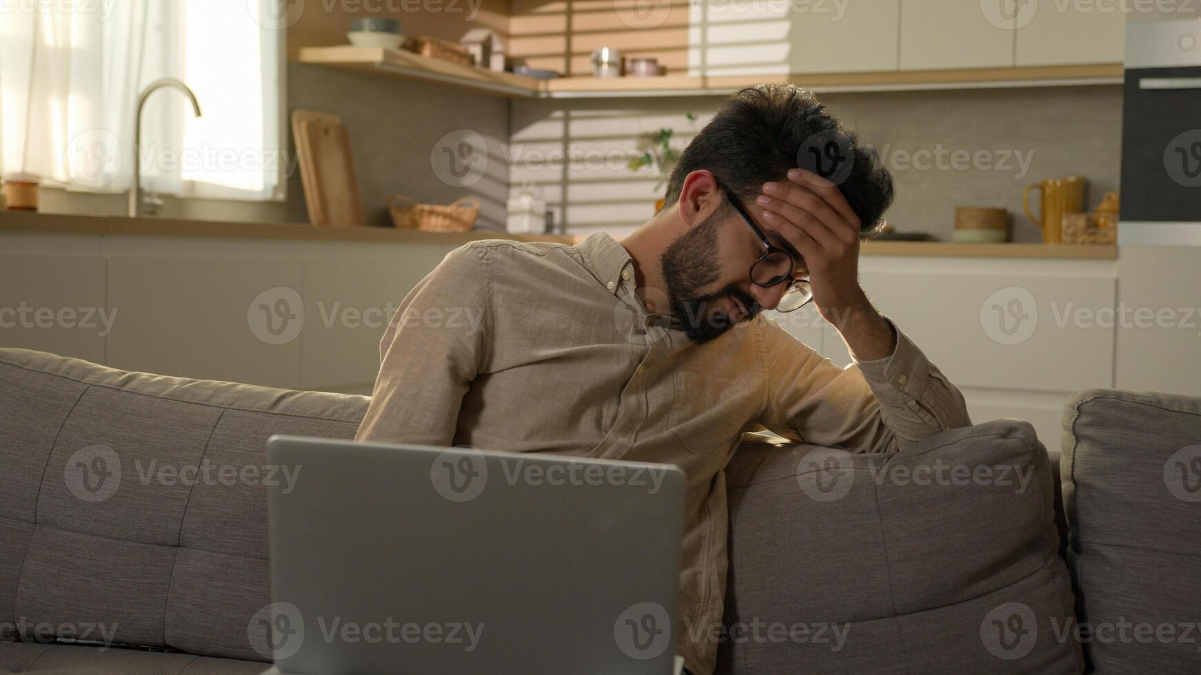 Shocked upset Indian Arabian man guy business work from home kitchen negative bad result worried anxious male freelancer on couch sofa read laptop notification computer problem fail difficult crisis photo