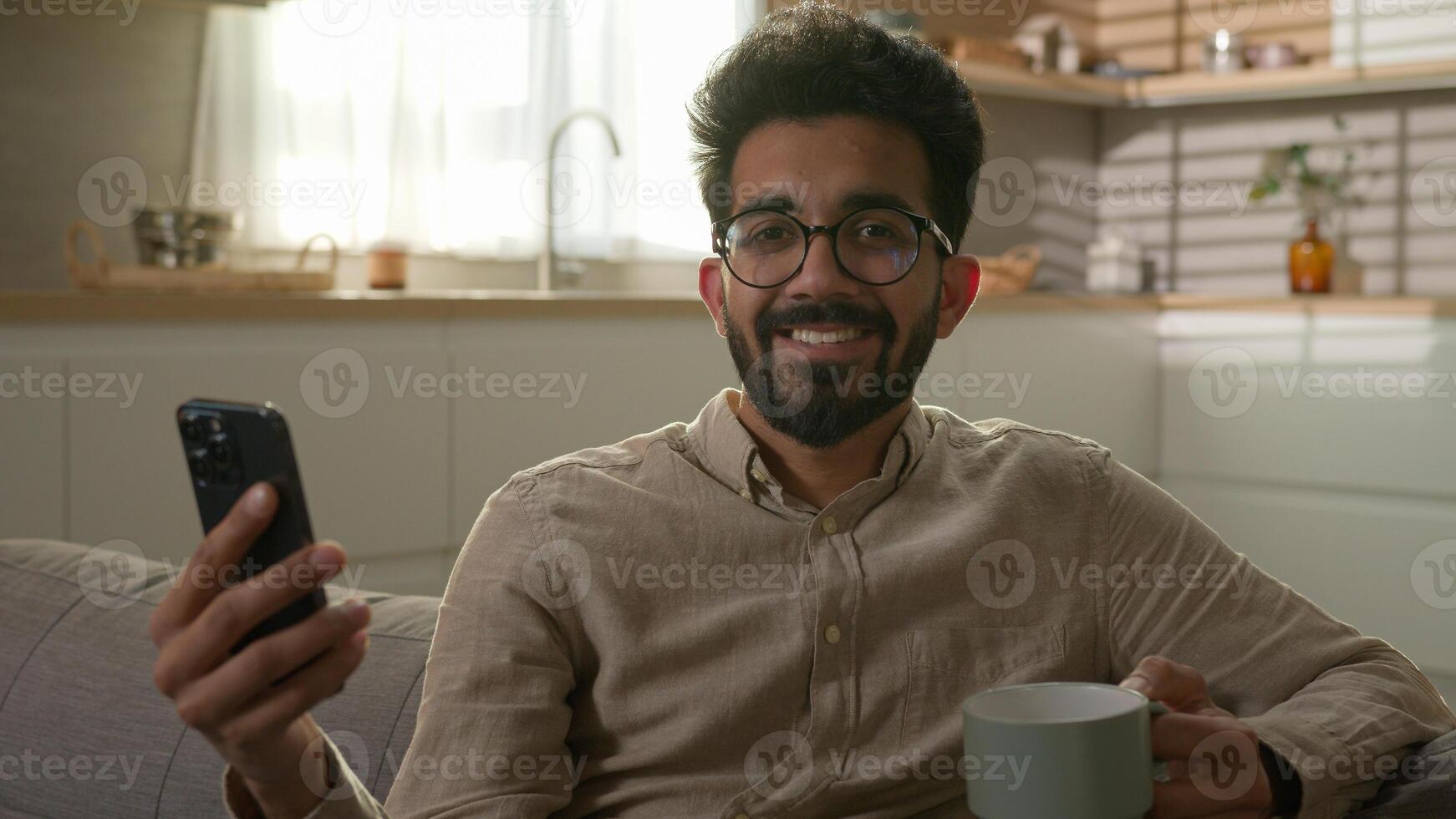Arabian Indian man ethnic homeowner male joyful happy guy in home kitchen watching online TV movie on mobile phone reading news drinking morning coffee relaxing on couch looking at camera smile photo