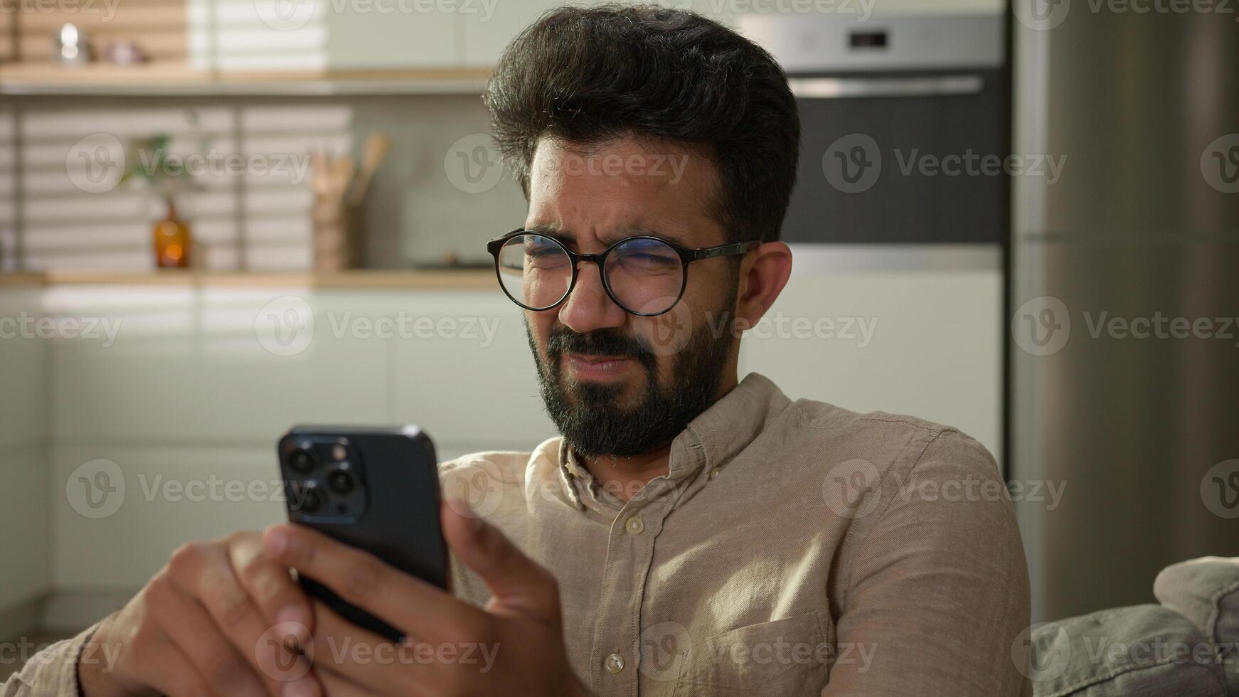 Confused dissatisfied Indian Arabian man guy on couch male homeowner at domestic kitchen using smartphone gadget read social media upset with bad news on mobile phone disgust unpleasant facial emotion photo
