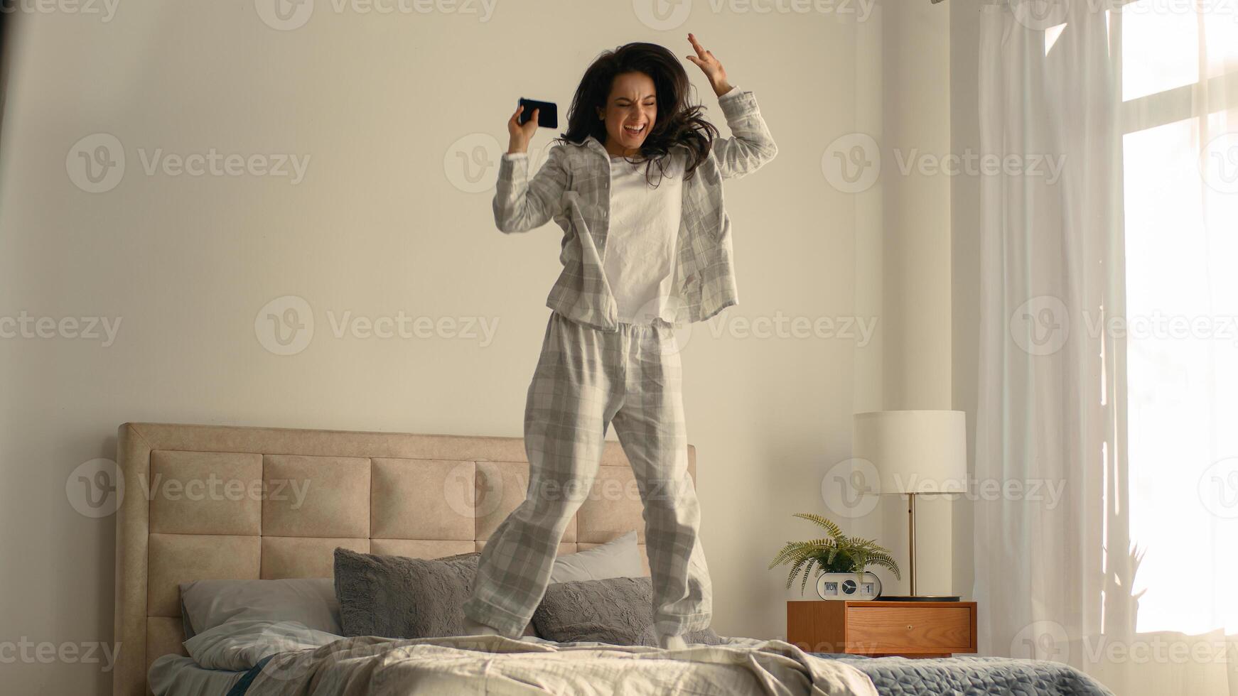 Young woman sitting on bed using mobile phone cellphone notification message good news home bedroom inside female rejoicing smiling laughing jumping fun luck victory success celebrate delighted gadget photo