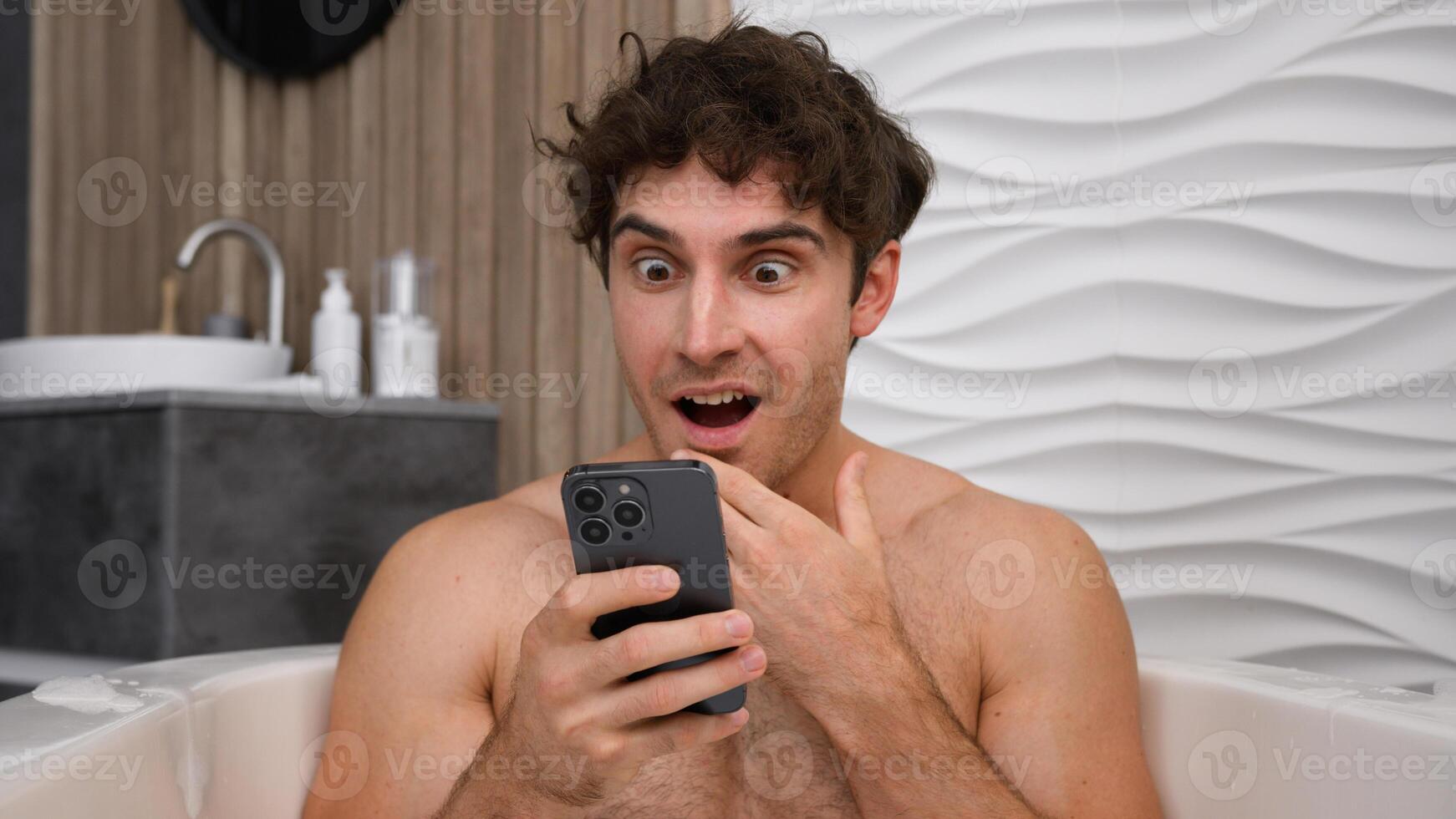 Caucasian man naked male guy relaxing in bath with soapy foam relax rest in bathroom bathtub amazed shocked surprised looking smartphone reading good news in mobile wow reaction astonish washing tub photo