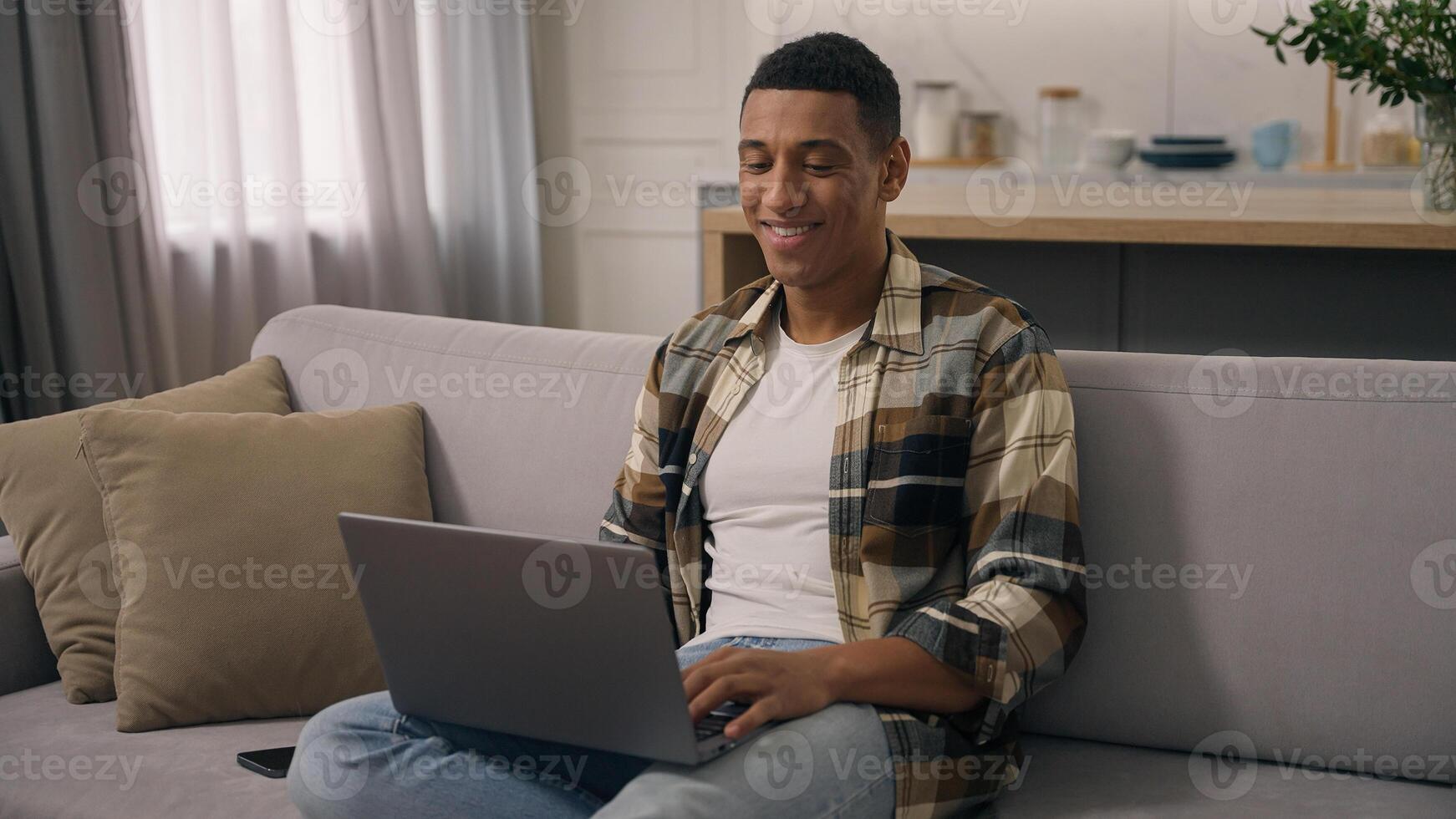 Happy smiling African American ethnic man working at home distant online work studying at sofa guy laptop using computer at couch male using pc playing internet game use app shopping chatting photo