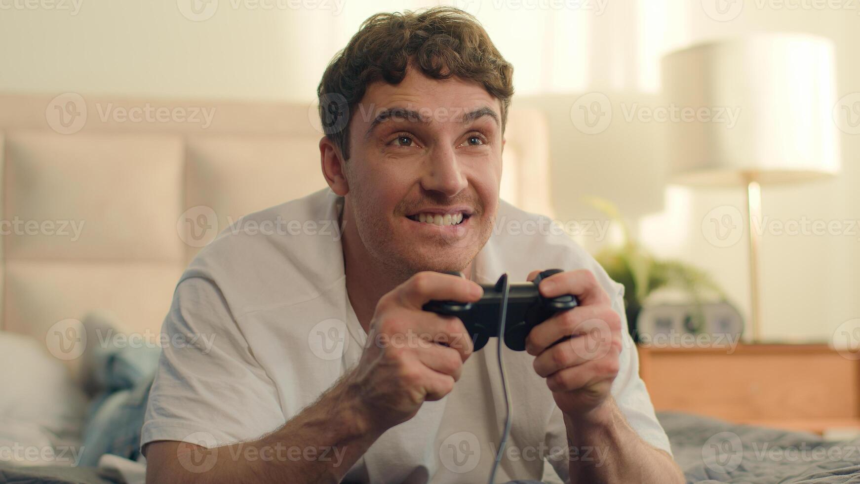 Playful Caucasian European young man lying on bed playing console holding joystick games fun weekend home enjoy guy hobby leisure hands technology virtual cyberspace cheerful excitement smiling photo