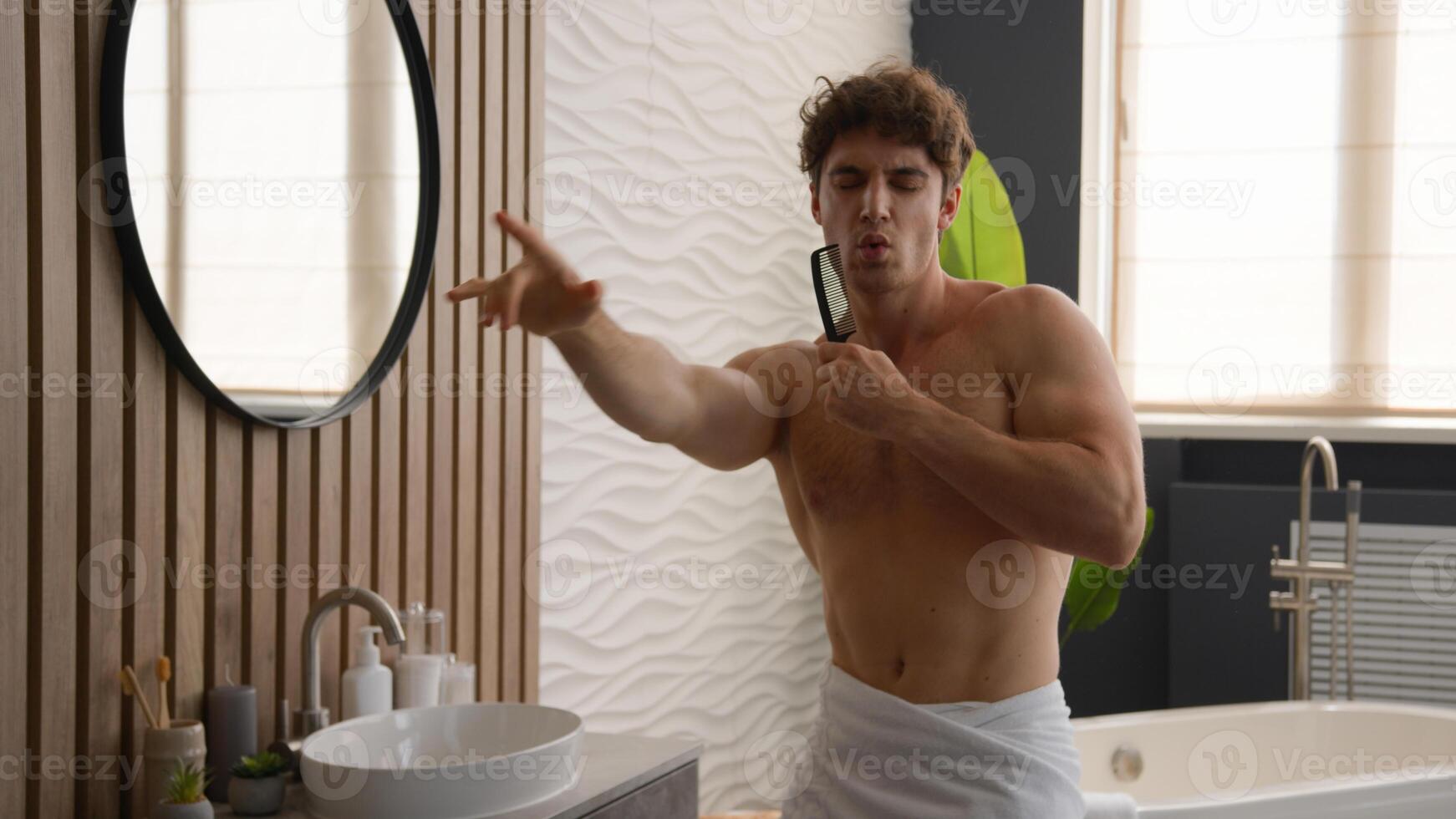 Joyful energetic enthusiastic funky naked Caucasian happy guy male singing song in hairbrush after brushing hair sing in comb brush looking mirror home hotel bath joy man in bathroom dancing fun music photo