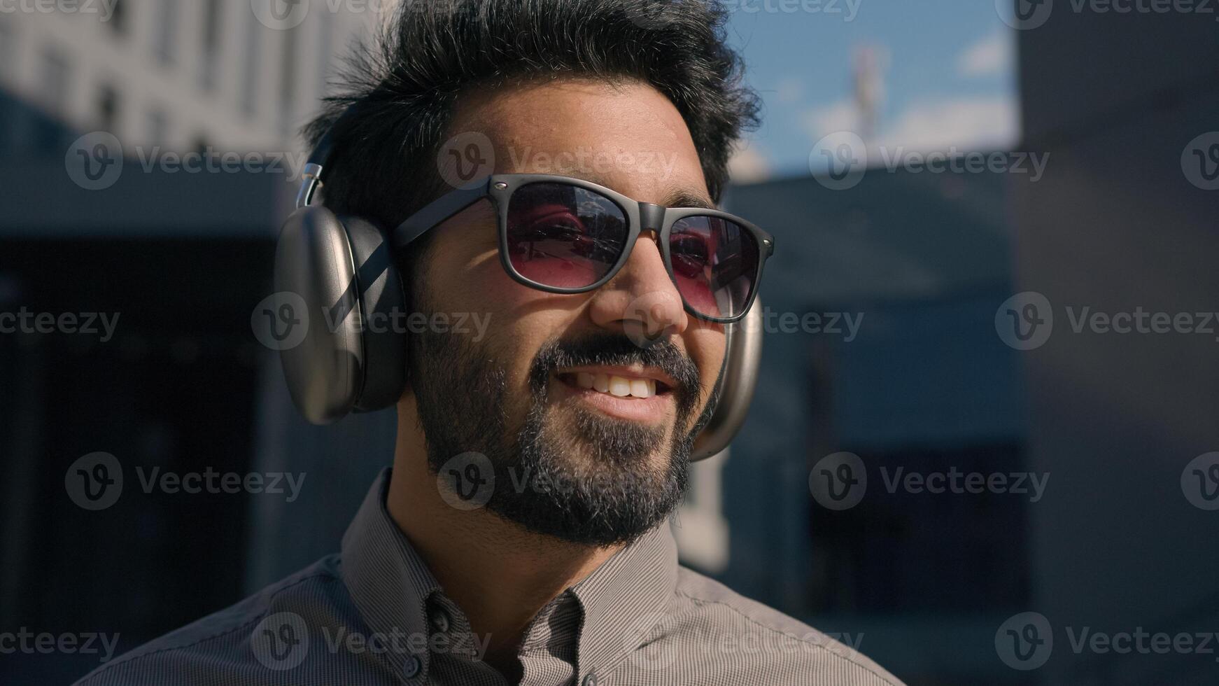 Carefree Indian Arabian businessman business man student wear sunglasses listen music sunny outdoors enjoy sound in wireless headphones near city office portrait relaxed happy guy smile in earphones photo