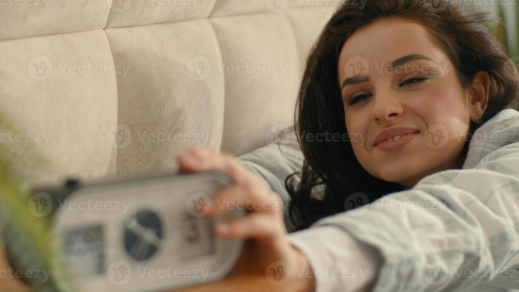 Happy satisfied woman lying in bed bedroom home comfortable cozy waking up alarm clock good morning smiling pleasure rest female girl tenant awake daydream joy relaxing weekend electronic technology photo