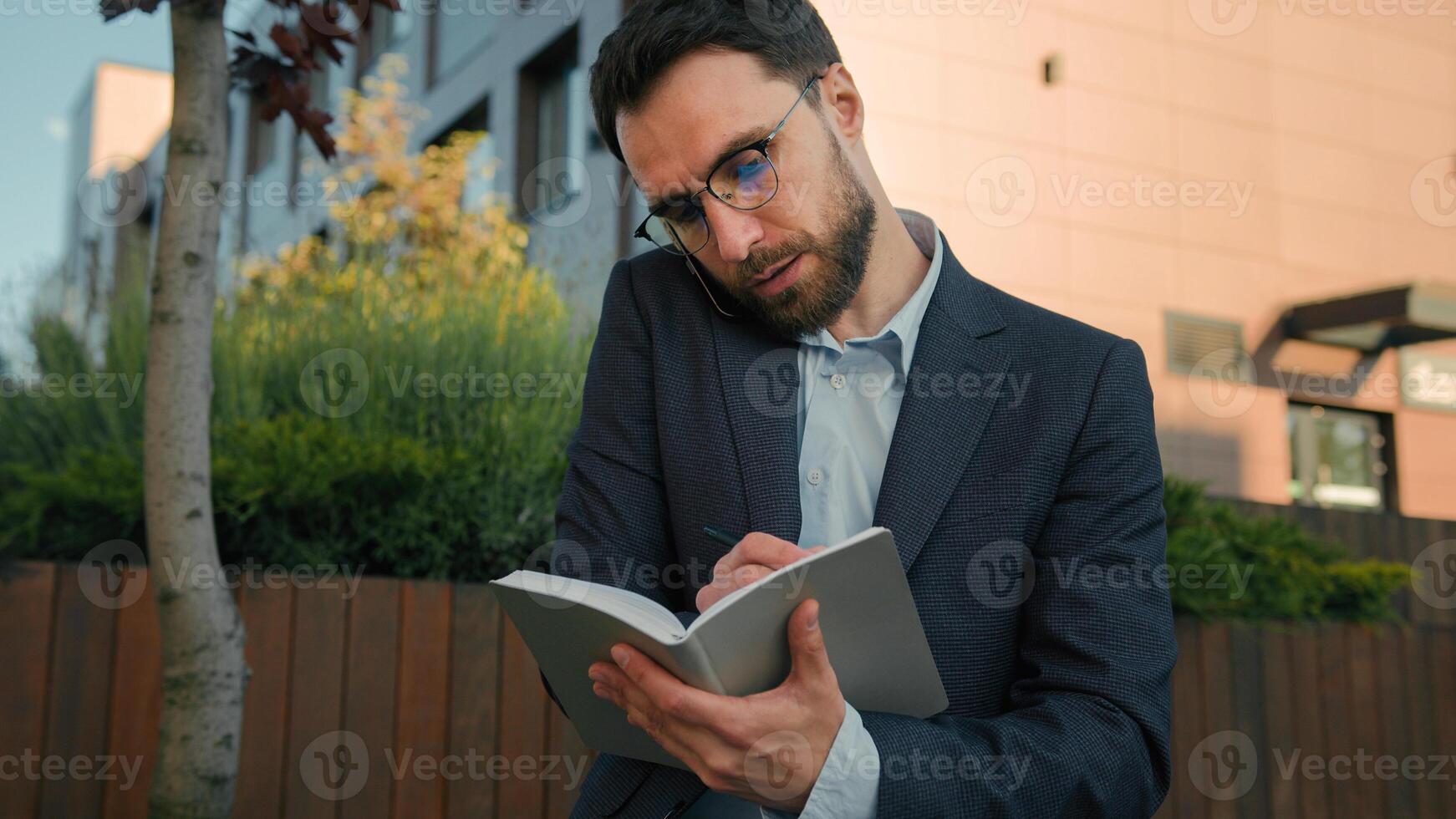 Busy Caucasian adult businessman talking with client mobile phone conversation writing notes multitasking man male manager working outdoors write schedule in notebook book business consulting in city photo