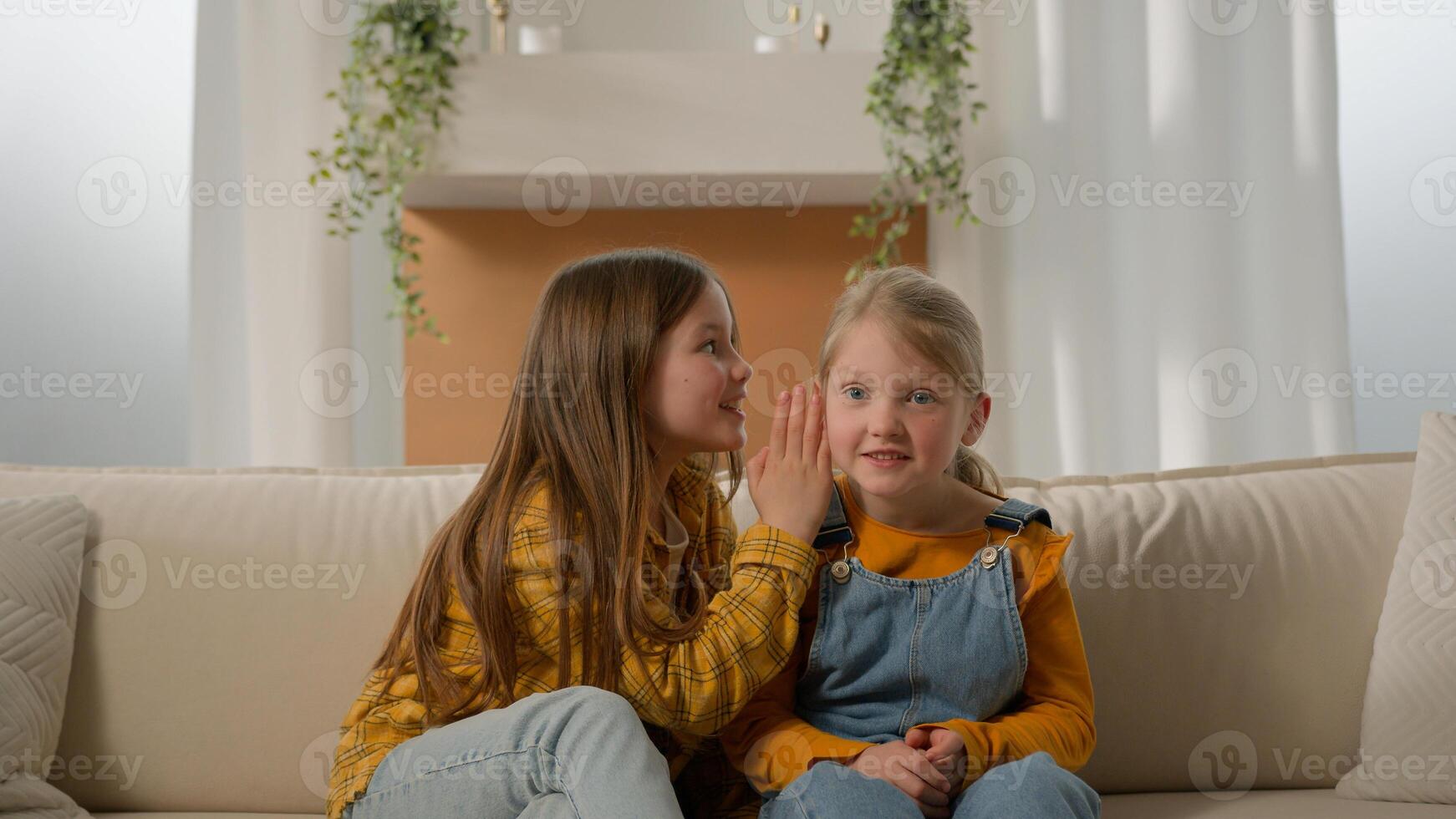 Naughty carefree Caucasian children little girls sisters kids talk whispering in ear rumors amusing story. Daughters siblings gossiping share news secret information together couch indoors friendship photo