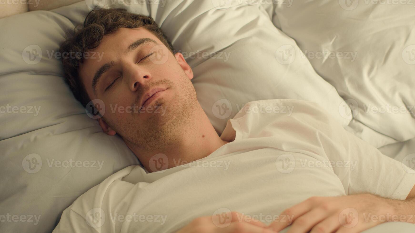Caucasian happy man sleeping open eyes wake up looking at camera smiling good morning comfortable cozy guy rest relaxation bedroom bed hotel wellbeing positive weekend male pleasure home satisfaction photo