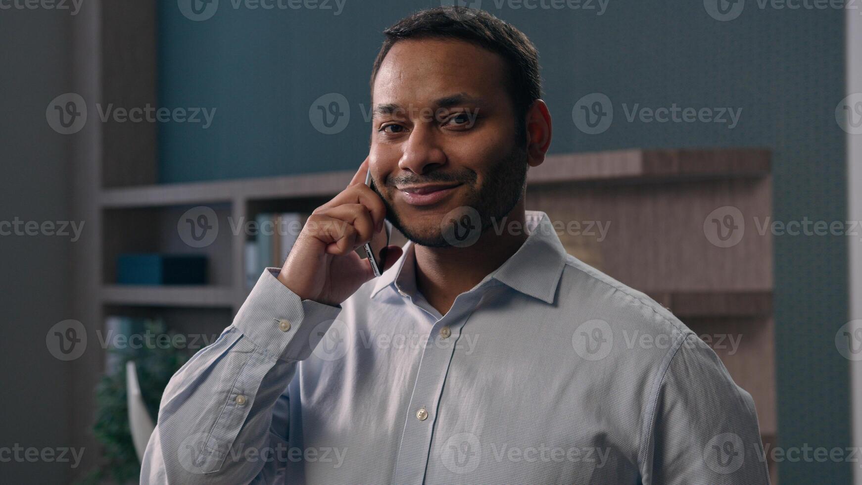 American male office worker at workplace in corporate company businessman talking mobile phone smiling multiracial man speak business negotiate discuss answer call smiling at camera talk smartphone photo