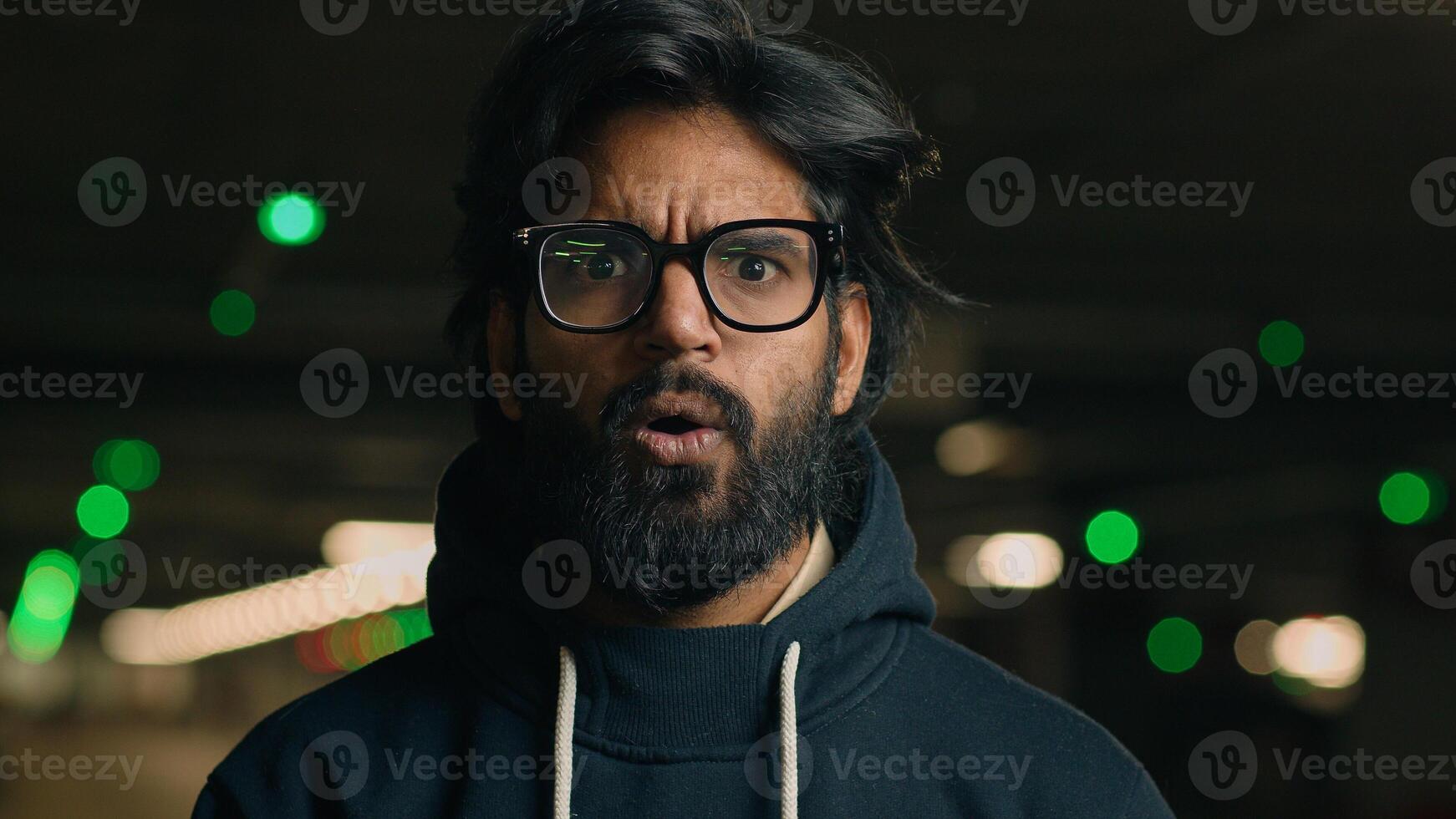 Close up Indian shocked bearded guy in glasses astonished looking at camera at dark underground wonder amazed Arabian man shock fear scared say wow stunned big eyes open mouth bad news at parking lot photo
